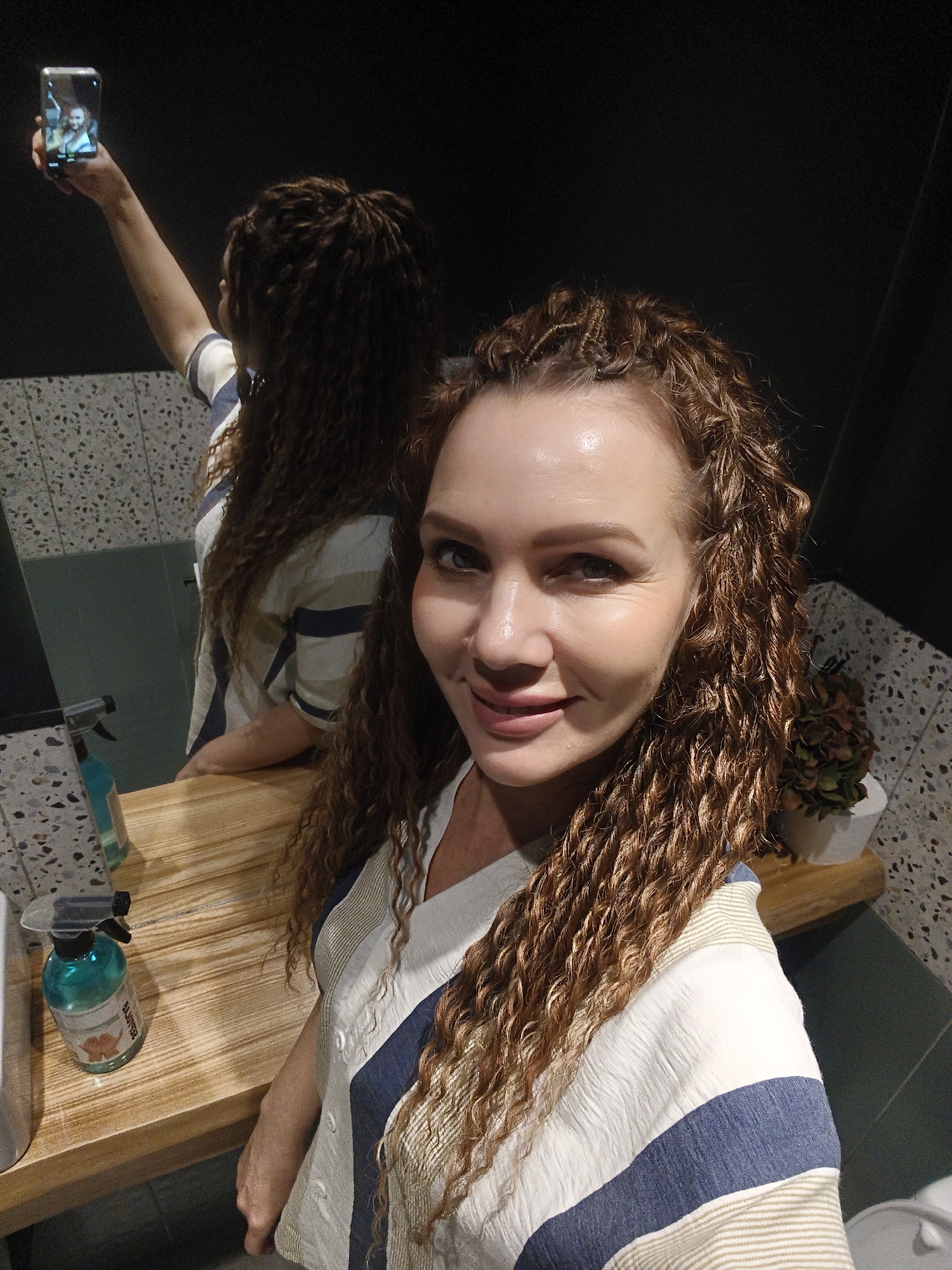 A woman with curly auburn hair and fair skin is taking a selfie in a bathroom. She is wearing a white and blue striped top. A mirror behind her reflects her taking the picture with her phone. On the counter, there is a wooden surface with a bottle of cleaning spray and a small potted plant. The walls are a mix of speckled gray and dark gray. Location is Unknown, Unknown.