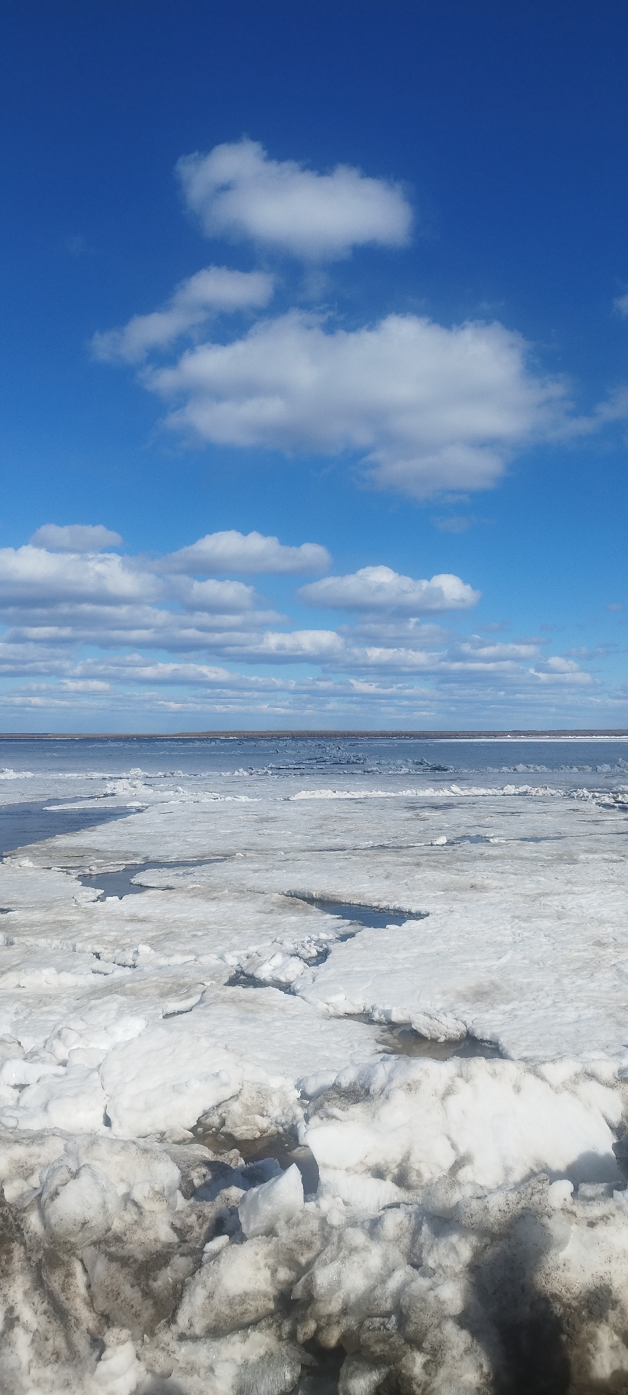 The image depicts a frigid, icy landscape with a partially frozen body of water under a bright, partly cloudy sky. The foreground is dominated by chunks of dirty, melting ice and snow piled together. Beyond the ice, a dark, open body of water stretches toward the horizon. The sky is a clear blue, dotted with white, puffy clouds. The location is likely in or near Yakutsk, Russia, considering the presence of the frozen river and the prevalence of cold conditions. There are no people visible. The overall scene suggests a transition between winter and spring, with the ice beginning to thaw.