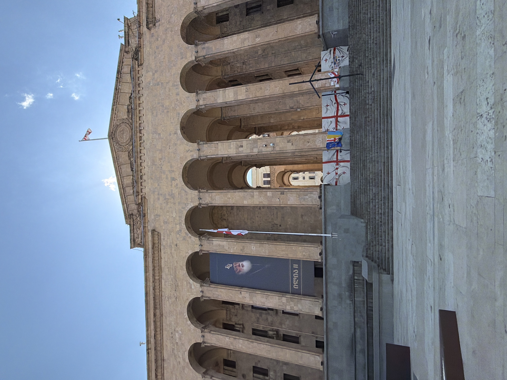 The image captures a worm's-eye view of a grand, light-colored stone building, likely a prominent public structure such as the Parliament of Georgia in Tbilisi, oriented vertically due to the original image's rotation. The building features multiple levels with deep arched loggias or recessed balconies, supported by robust columns, creating a sense of classical or neo-classical architecture. Its facade is adorned with intricate carvings and decorative elements, particularly noticeable near the roofline.
Bright sunlight illuminates the scene, casting soft shadows and indicating daytime. The sky is a clear, vibrant blue with a few scattered white clouds.
Several flags are visible. A small red and white flag, characteristic of the Georgian national flag (St. George's Cross), flies from a pole at the highest point of the building's pediment. Further down, multiple large white banners emblazoned with red crosses—also representing the Georgian flag—are hung from poles along the building's side. Among these, a smaller Ukrainian flag (blue and yellow) is prominently displayed, symbolizing solidarity.
A large vertical banner hangs from one of the arched sections on the lower part of the building. It features a solemn portrait of an elderly man with a white beard, wearing religious vestments and a miter adorned with a cross, identifying him as a high-ranking cleric. Text in Georgian script accompanies the portrait, which reads "ილია II" (Ilia II), indicating Catholicos-Patriarch Ilia II of Georgia.
The foreground and right side of the image show a wide, light-colored paved area, suggesting a public square or plaza leading up to the building. The overall impression is one of national pride and possibly a commemoration, set against the backdrop of an important architectural landmark in Tbilisi, Georgia.