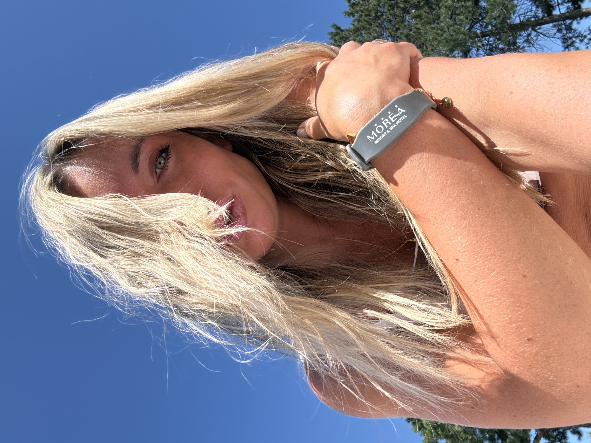 The image is a low-angle, close-up shot of a woman with long, blonde hair against a bright blue sky. Her hair is partially covering her face, obscuring her nose and mouth, but her left eye is visible, looking towards the camera. She is wearing a grey wristband with the text "MOREA RESORT & SPA HOTEL" printed on it, and a small gold charm hangs from it. Her right arm is bent, and her hand is resting on her arm. The background shows the tops of green trees and the clear blue sky. The lighting suggests it is daytime and sunny. The image is an outdoor portrait taken in the city of Anapa, Russia.