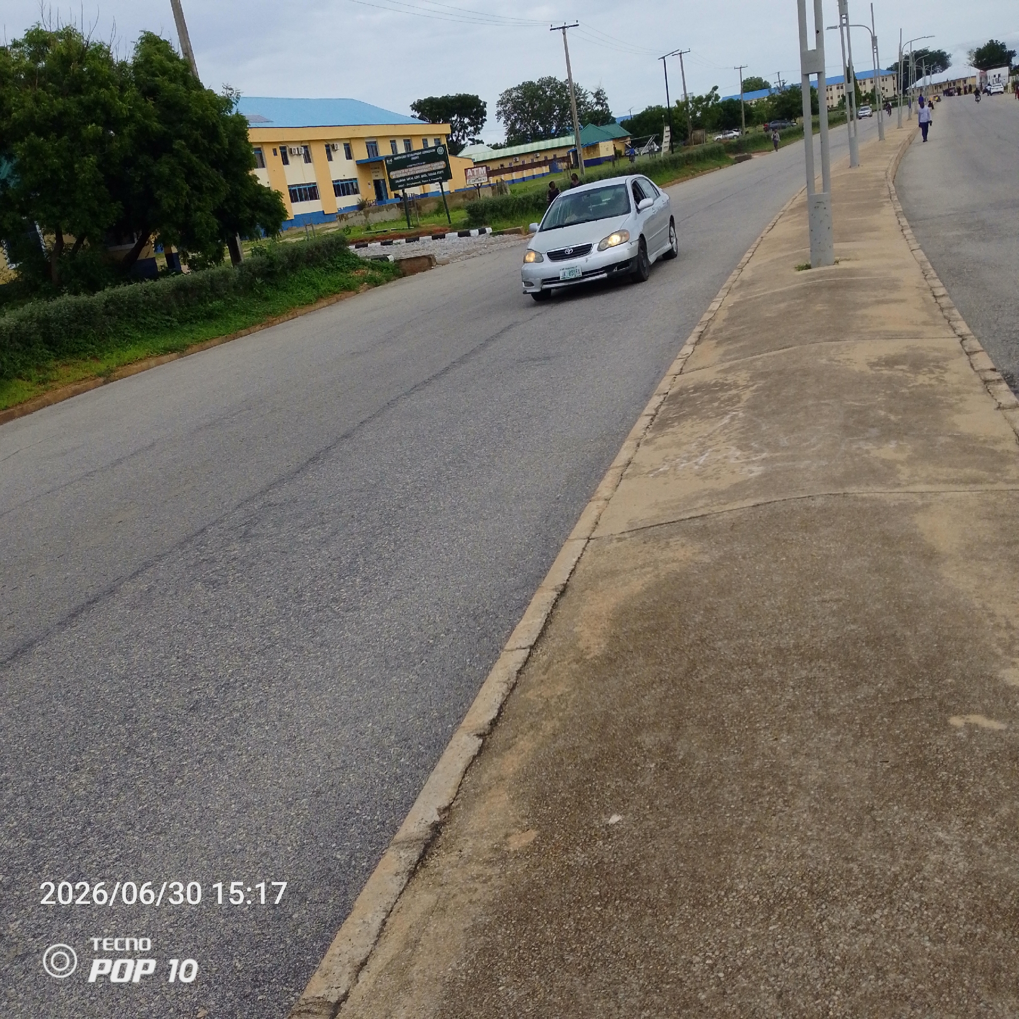 A silver sedan drives along a paved road in Jalingo, Nigeria. The car is heading towards the right side of the frame and appears to be the main focus of the image. In the background, a long, yellow building with a blue roof stands prominently, suggesting it might be an educational institution or government building. Other smaller buildings and trees are visible further down the road. The sky is overcast, indicating a cloudy day. Several streetlights line the right side of the road, bordering a concrete walkway. A few people are scattered along the road and sidewalk, engaged in various activities like walking or standing. The timestamp "2026/06/30 15:17" and "TECNO POP 10" are overlaid on the bottom left of the image.
