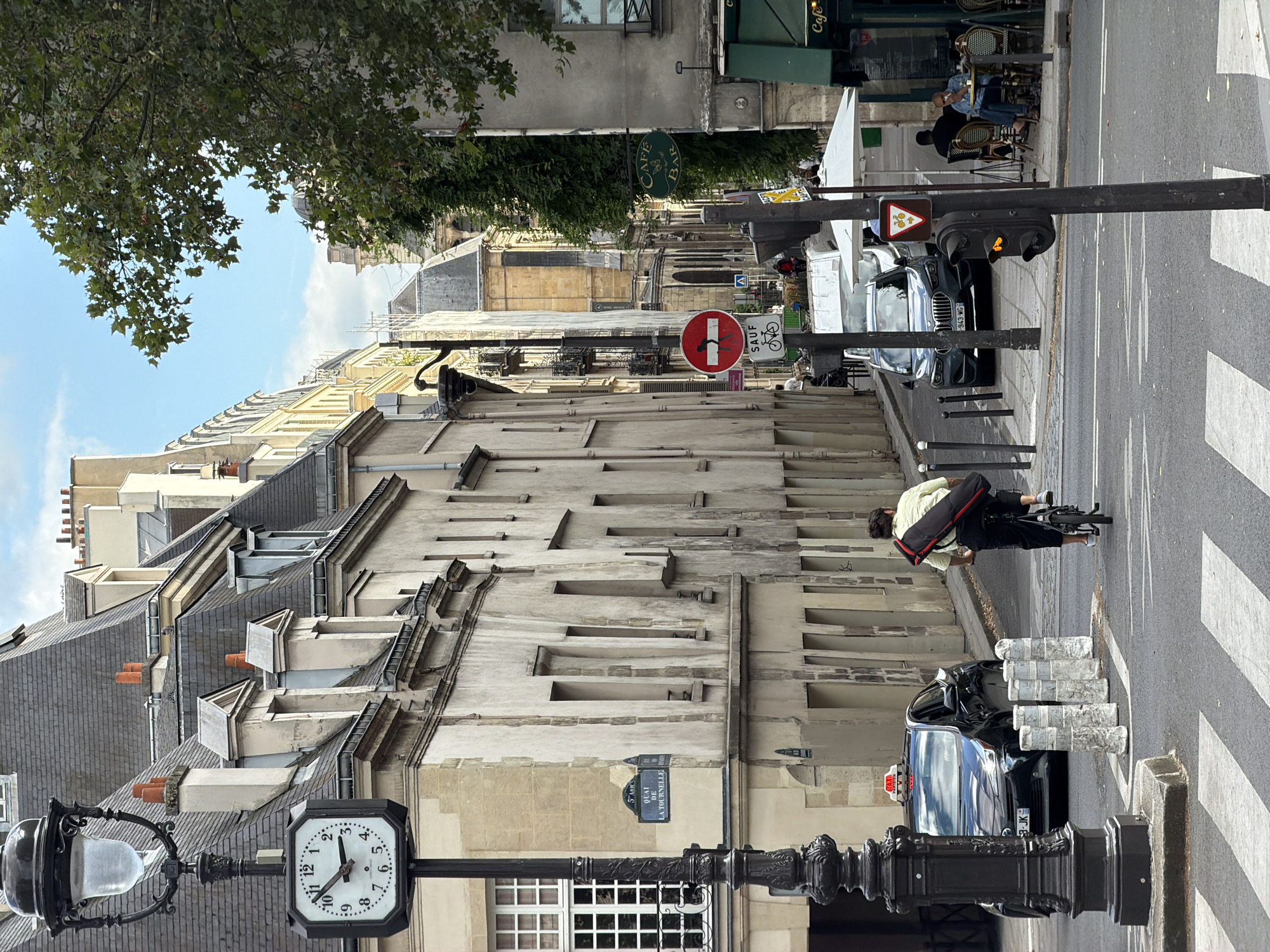 This is a vertical, eye-level shot of a Parisian street. The scene is captured during the day, with clear skies suggesting fair weather. In the foreground, a traditional Parisian lamppost with an ornate base and a frosted glass lantern stands to the left. To its right, a clock face shows the time as approximately 10:03.
The street is lined with classic Haussmannian buildings, characterized by their stone facades, mansard roofs, and numerous dormer windows. The architecture suggests a historic and established urban environment.
Several people are present. A person on a bicycle, wearing a light-colored shirt and dark pants, is moving away from the camera. In the background, patrons are seated at tables outside a "Cafe Bar," enjoying beverages. A taxi is parked on the street.
Visible signage includes street name signs that read "QUAI DE LA TOURNELLE" and "5 ARR." Also present are traffic signals and various street signs, including a "No Entry" sign with a pictogram of a person and a bicycle, and a pedestrian crossing marked with white stripes on the asphalt. The overall atmosphere is that of a typical, bustling Parisian street scene.
The crops highlight details such as the leaves of a tree in the upper left, the detailed architecture of the buildings, the signage, and the various elements of street furniture and traffic control.