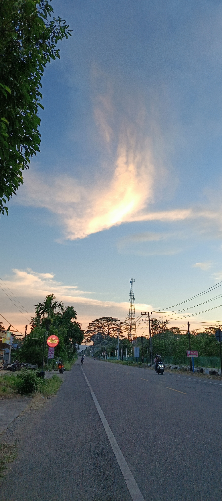 This is a vertical, eye-level, medium shot of a paved road in Sukamulia, Indonesia at dusk. The sky is a soft blue with a prominent cloud formation illuminated by the setting sun, casting a warm, peachy glow. To the left of the frame, a lush green tree canopy partially obscures the sky. The road, marked with a white center line, stretches into the distance, flanked by vegetation and a few structures. A tall telecommunications tower stands prominently on the right side of the road, its metal lattice structure silhouetted against the colorful sky. A few motorcycles are visible on the road, with one rider in the foreground and two in the middle distance. The overall scene depicts a peaceful, rural-industrial landscape transitioning from day to night.