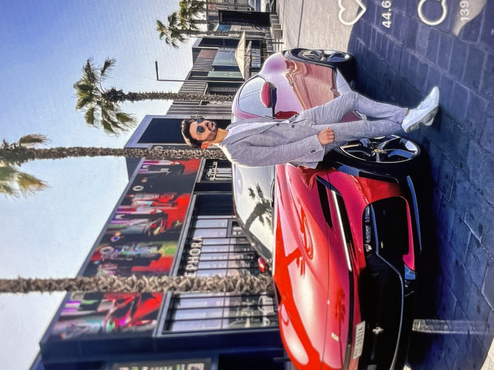 A man in a light grey suit and white sneakers stands next to a bright red Ferrari. He is leaning against the car, with his right hand on the vehicle. He wears sunglasses and has a beard. The car is parked on a paved area in front of a building with large windows and a sign that reads "SEPHORA". Palm trees line the street. The sky is blue and clear.