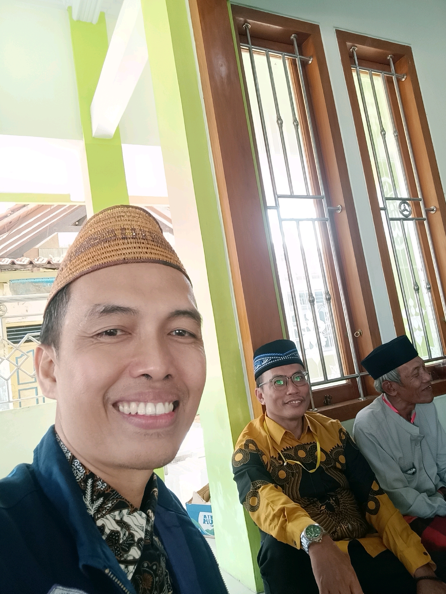 The image is a selfie taken by a man in the foreground, smiling at the camera. He is wearing a woven, conical hat and a dark blue jacket over a patterned shirt. To his right, two other men are visible. The man in the middle is wearing a yellow and black patterned shirt and a black cap with blue trim. He is wearing glasses and a watch. The man on the far right is wearing a grey shirt and a black cap. They are sitting in what appears to be an indoor waiting area or common space. The background shows wooden window frames with metal grilles and a light green pillar. The setting is identified as Penusupan, Indonesia.