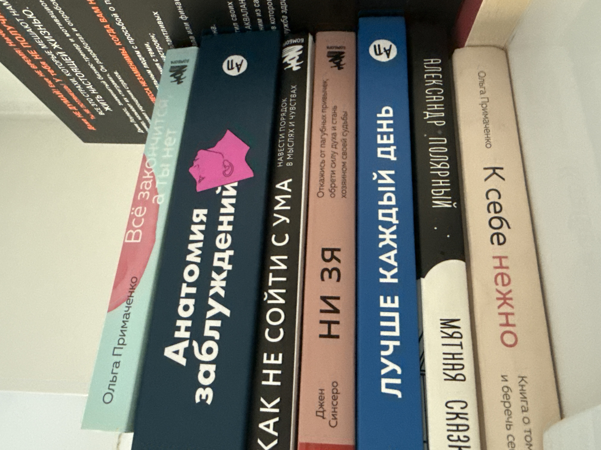 The image shows a stack of books on a shelf. The books are in Russian, and the titles are partially visible. The spine of the top book is light blue with pink text. The second book from the top is dark blue with white text that reads "Анатомия заблуждений". The third book from the top is black with white text that reads "КАК НЕ СОЙТИ С УМА". The fourth book is pink with black text that reads "Джен Синсеро НИ ЗЯ". The fifth book is dark blue with white text that reads "ЛУЧШЕ КАЖДЫЙ ДЕНЬ". The sixth book is white with black text that is upside down and appears to be "МАТНАЯ СКАЗК". The seventh book is light pink with red text that appears to be "К себе лично". The bottom of the image shows a white surface, possibly a shelf or table. There is no information in the image to suggest the location is Chișinău, Moldova.
