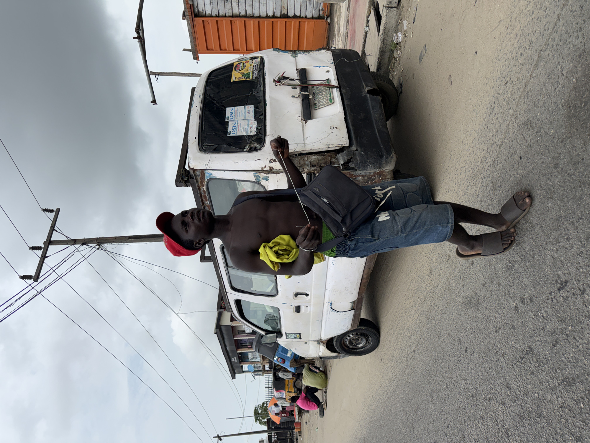 A shirtless man in a red cap and blue denim shorts stands in front of a dilapidated white van, holding a yellow cloth. He has a black shoulder bag and wears sandals. The van is parked on a paved street in Ado, Nigeria, near a building with an orange upper section. Power lines and poles are visible overhead against a cloudy sky. In the background, a small crowd of people gathers near other vehicles.