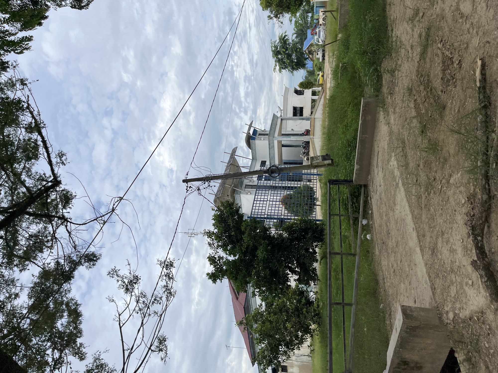The image depicts a building in Bundar, Indonesia, under a cloudy sky. The building is modern in design, with white walls, blue accents, and a red tiled roof. It appears to be a commercial or institutional building, possibly an office or a small factory, with a parking area in front. Several vehicles, including motorcycles, a yellow car, a white truck, and a tow truck with a crane, are parked in the vicinity. The surrounding area is covered in green grass and trees, with a dirt path leading towards the building. There is a metal fence in the foreground, and a concrete barrier. The overall scene suggests a functional, developed area within the city.