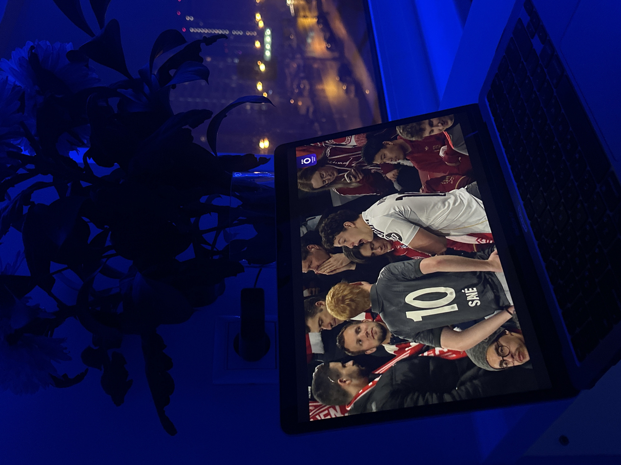 A laptop sits on a surface, displaying a soccer match, presumably in Moscow, Russia. The laptop is positioned on a table, and the screen shows several soccer players in close proximity. On the left is a potted plant, and through the window behind the laptop, city lights of Moscow are visible. The overall lighting is a blue hue, potentially from an artificial light source.