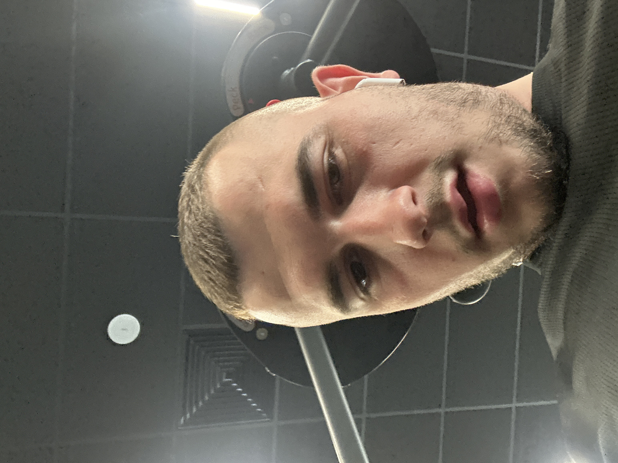 A close-up selfie photograph of a young man with short, light brown hair and a trimmed beard. He appears to be at a gym, as suggested by the presence of exercise equipment partially visible in the background. The man is wearing an earbud in his right ear. The background features a dark gray ceiling with visible ventilation and a light fixture. The image does not contain any contextual elements to determine whether or not it was taken in the city of Plovdiv, Bulgaria.