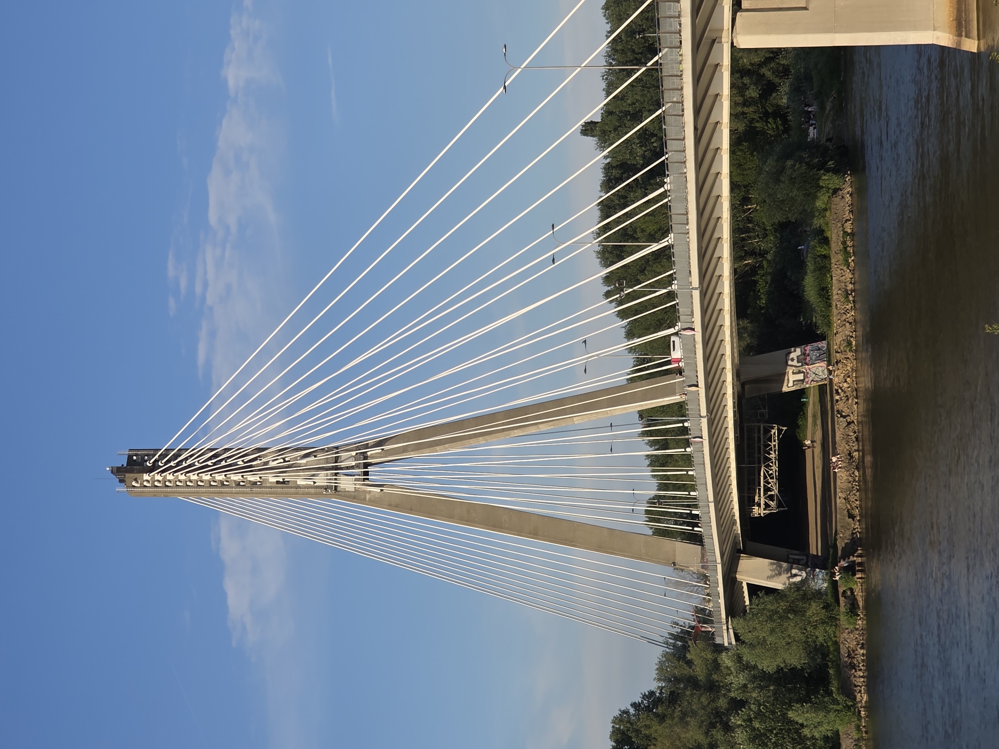 This is a vertical, eye-level, full shot of the Świętokrzyski Bridge in Warsaw, Poland, on a clear day with a blue sky and scattered clouds. The bridge, a cable-stayed structure, dominates the frame with its two concrete pylons and numerous white cables. The roadway of the bridge is visible, with cars and lampposts. Lush green trees line the riverbank on both sides. The Vistula River flows in the foreground, its surface rippled by the current. Several people are visible on the riverbank beneath the bridge, some sitting and others standing. The bridge pylons are concrete, with the main pylon having a tall, rectangular structure at its apex. There is graffiti visible on the concrete support structures under the bridge. The sunlight suggests it is late afternoon.