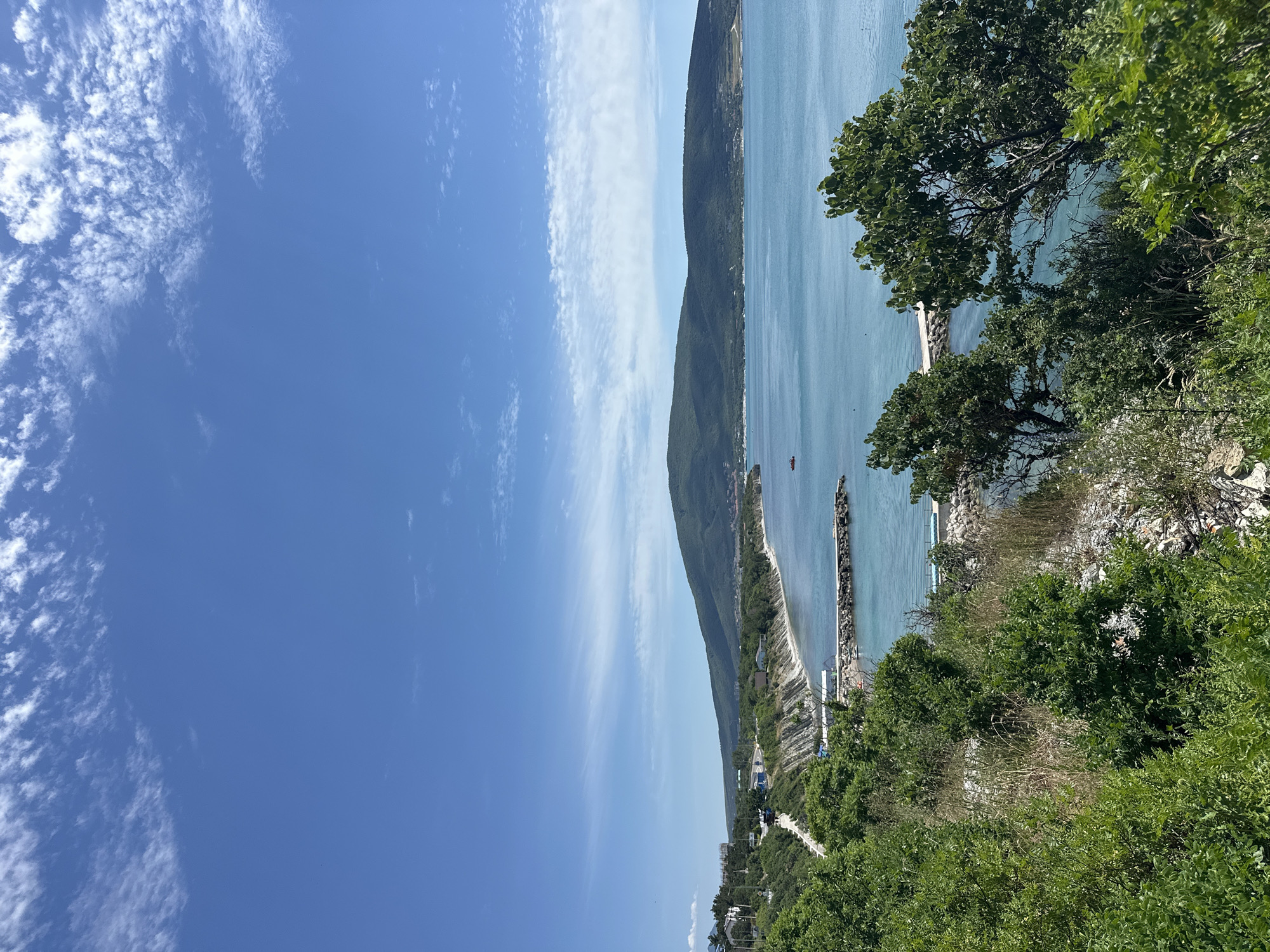 The image captures a scenic view of the Black Sea coast in Kabardinka, Russia. In the foreground, lush green vegetation covers a hillside, with glimpses of rocky terrain. A paved road winds its way along the cliffside, bordered by more greenery and some scattered buildings. The middle ground features the calm, turquoise waters of the sea, with several breakwaters extending into the water. A small red boat is visible in the distance on the sea. The coastline curves around, with a low, forested mountain range dominating the background. The sky is a vibrant blue, with scattered white clouds creating a pleasant summer atmosphere. The overall scene depicts a serene coastal landscape with elements of human infrastructure integrated into the natural environment.