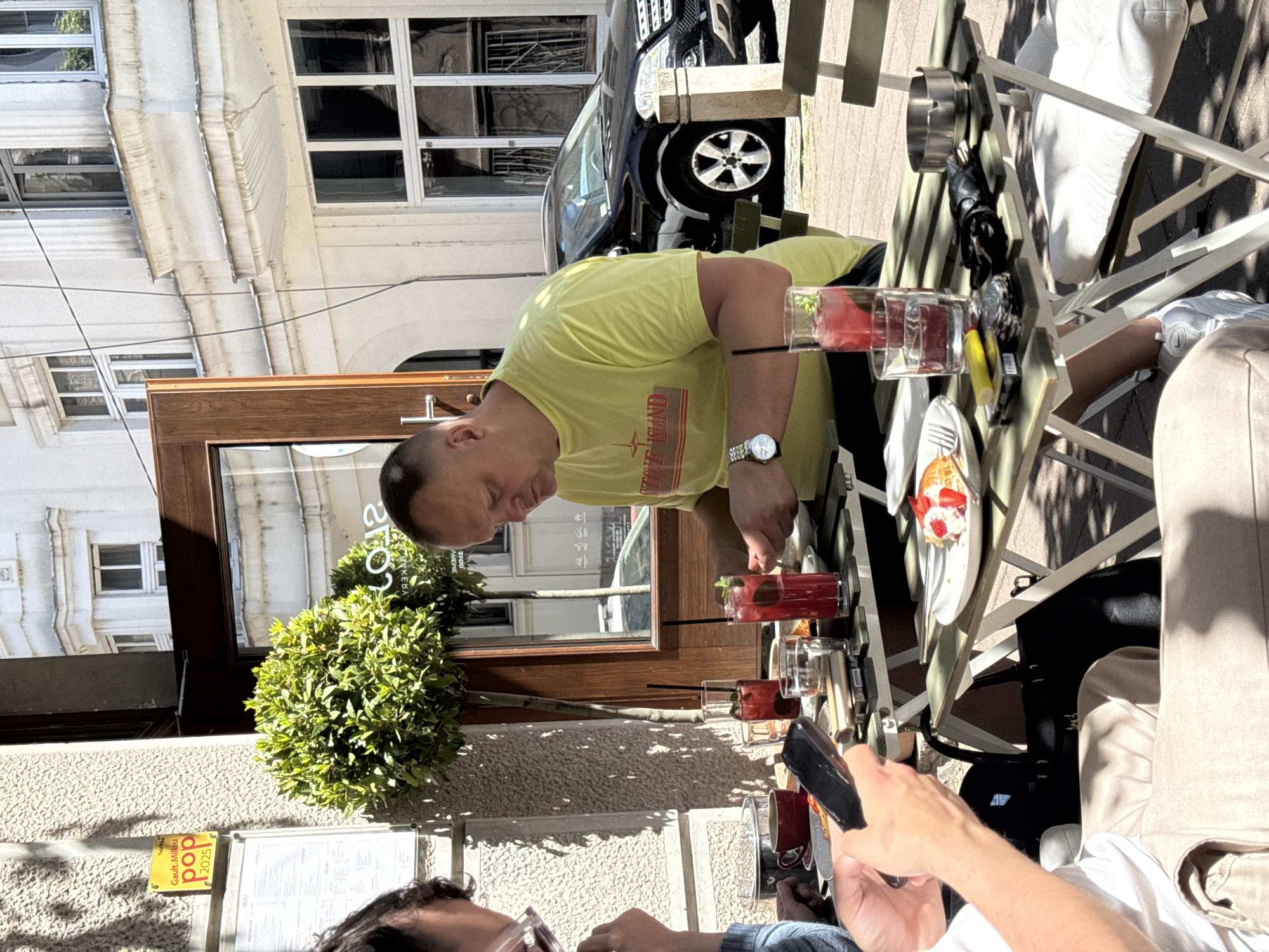 The image depicts a group of people dining outdoors at a café in Belgrade, Serbia. One person, wearing a yellow shirt with a red logo, is leaning over the table which has several glasses of red drinks, a plate with a dessert, and other small items. A second person is holding a cell phone, possibly taking a photograph of the drinks. The café appears to be located next to a building with a dark wooden door, and a menu. A tree is next to the café. In the background, a black car is parked on the street. The image is taken during the daytime, and the scene is well-lit by sunlight.