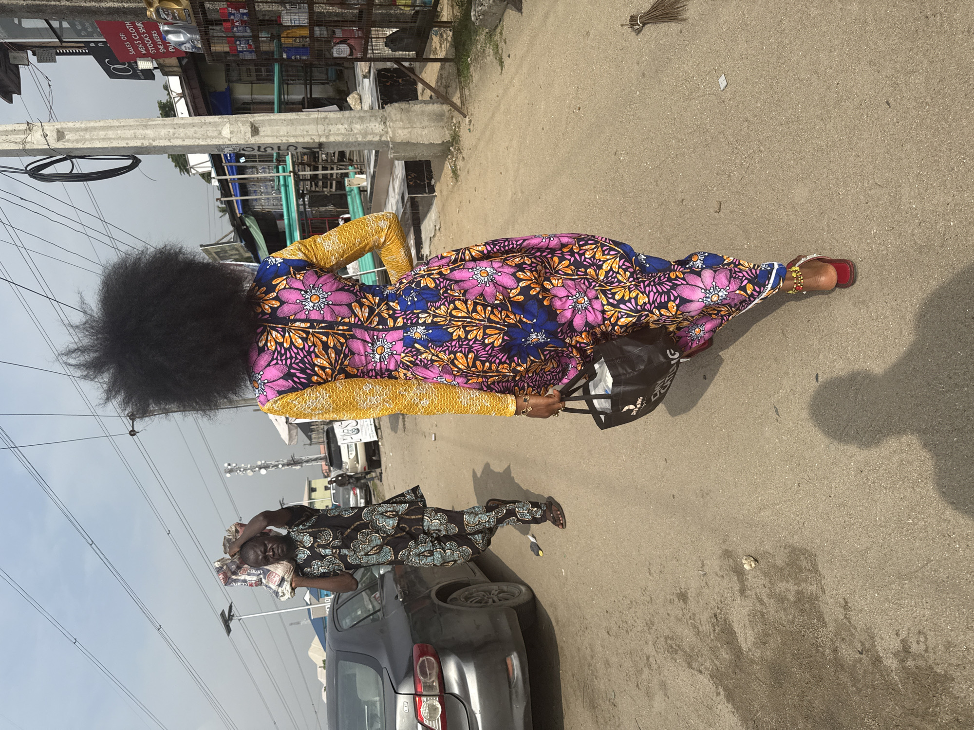 The image, rotated 90 degrees, captures a vibrant street scene in an urban or semi-urban area, consistent with Ado, Nigeria, during bright daylight hours under a clear sky.
The primary subject is a woman walking away from the viewer, characterized by her voluminous, dark, curly afro hairstyle. She wears a striking, full-length dress featuring a rich pattern of large pink/magenta flowers with yellow centers, set against dark blue leaves and orange accents on a dark background. Her long sleeves are a contrasting bright yellow fabric with a subtle textured pattern. She sports red sandals adorned with beaded ankle bracelets, one yellow and one red. In her left hand, she carries a black tote bag with white lettering that is not fully legible.
Further down the street, on the left, another person is visible, possibly male, wearing patterned clothing with light blue/green designs on a dark background. This individual appears to be carrying a bundled item on their head and is looking towards the camera. A dark-colored car is partially visible parked or moving alongside this person.
The street is paved, and shadows suggest a sunny day. Utility poles with numerous overhead electrical or communication wires crisscross the sky. In the background, various buildings, suggesting shops or stalls, line the street. A red sign is partially visible, showing text such as "SALES OF MEN'S CLOTH," "STOCKS SHIRT," and "SPEAKERS." Another blurry sign indicates "AUTO SELL" and "USED." A traditional broom lies on the ground in the midground, and small pieces of litter are scattered on the roadside. The overall impression is one of everyday life and commerce in a bustling street environment.