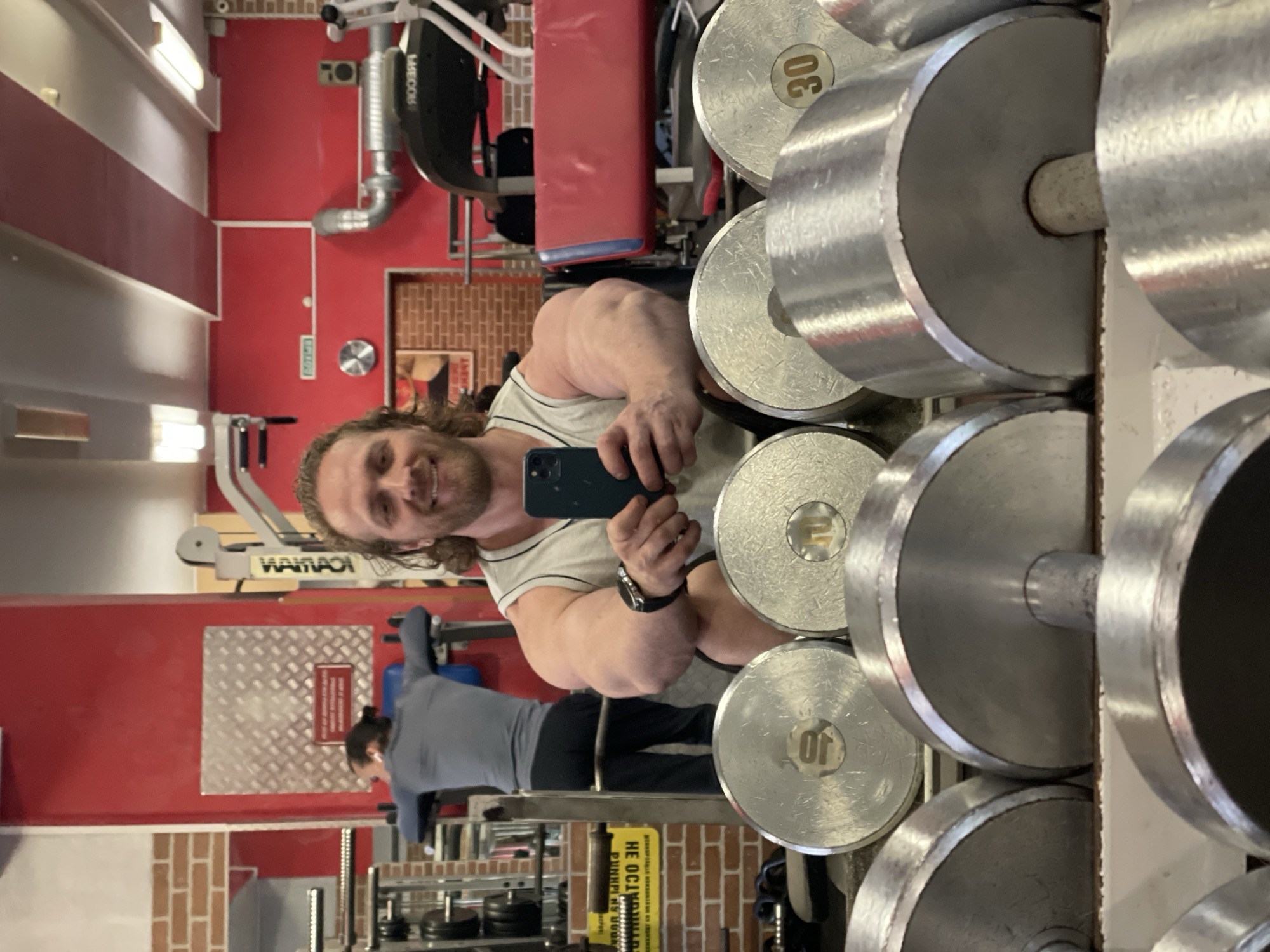 This is a portrait-oriented, vertically mirrored image taken in a gym. The primary subject is a man with long, wavy brown hair and a beard, smiling and holding a smartphone to take a selfie. He is wearing a light-colored tank top and a watch on his left wrist. His left arm is visibly muscular.
In the background, a rack of dumbbells is prominently displayed. Some of the dumbbells have weight markings, including "30" and "10". To the left of the man, a red wall dominates the scene. A piece of gym equipment, possibly a weight machine, is visible with the brand name "WAHHAO" partially visible. Further in the background, another person in a grey athletic shirt and black pants is working out, mirrored in a reflective surface.
There are several textual elements visible in the image. The weights on the dumbbells are marked with numbers. The gym equipment has a partial brand name. A yellow sign with Russian text is visible on the wall behind the person working out, which appears to read "НЕ ОСТАНАВЛИВАЙСЯ" (Don't stop). Another red sign with white text is on the wall. The overall setting is a gym, suggested by the equipment and the workout attire of the subjects. The lighting suggests it is indoors during the daytime. The man's smile conveys a sense of happiness or satisfaction. The mirrored nature of the image, along with the gym setting, suggests the man is taking a mirror selfie while at the gym. The location is identified as Saint Petersburg, Russia.