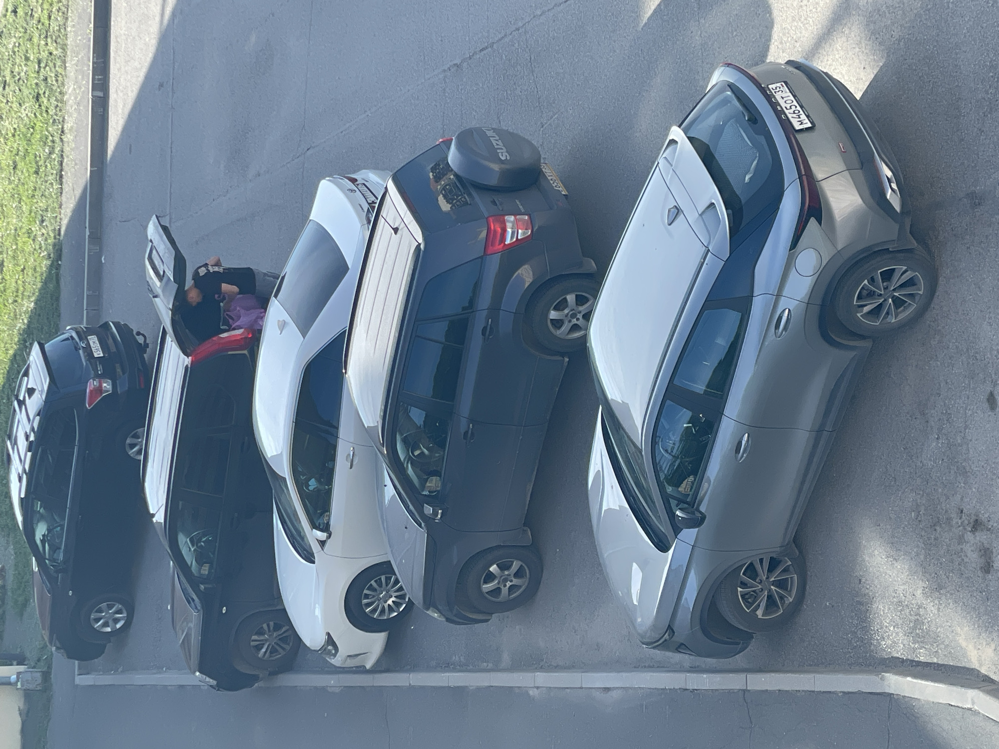 The image depicts an overhead view of five vehicles parked sequentially along a curb on an asphalt surface in the city of Vologda, Russia. The scene is illuminated by natural sunlight, casting shadows indicative of morning or late afternoon. A grassy area with foliage is visible in the background, beyond a sidewalk.
From top to bottom, the parked vehicles are:
1. A dark blue or black Subaru Forester SUV, equipped with roof rails. Its rear license plate is K565OK 35.
2. A black Nissan X-Trail SUV with its rear tailgate open. A person, dressed in a dark t-shirt with a light-colored graphic on the back, is visible standing at the rear of the vehicle, leaning into the open cargo area. A purple bag or item is partially visible within the trunk, suggesting loading or unloading activity. The license plate is not fully discernible.
3. A white Toyota Camry sedan. Its license plate is partially obscured and unreadable.
4. A dark grey Suzuki Grand Vitara SUV, distinguished by a rear-mounted spare tire cover bearing the "SUZUKI" brand. Its rear license plate is O565XT 35.
5. A grey OMODA C5 crossover SUV. Its rear license plate is M465OT 35.
All visible Russian license plates (K565OK 35, O565XT 35, M465OT 35) display the regional code "35," which corresponds to Vologda Oblast. The vehicles are parked parallel to a light-colored concrete curb delineating the parking area from a paved sidewalk.
