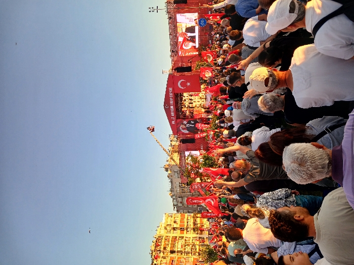 The image shows a large crowd of people gathered in a public square in İzmir, Türkiye, at the coordinates 38.436, 27.141. The crowd is facing a large stage with a screen and several flags hanging above it. A man is giving a speech from the stage, facing the crowd. The people in the crowd are listening intently and waving Turkish flags. The square is surrounded by buildings. The sky is blue and clear, with a few birds flying overhead. The scene is bright and lively, with a festive atmosphere. Many people are holding red and white flags and signs, all facing the stage and the speaker.