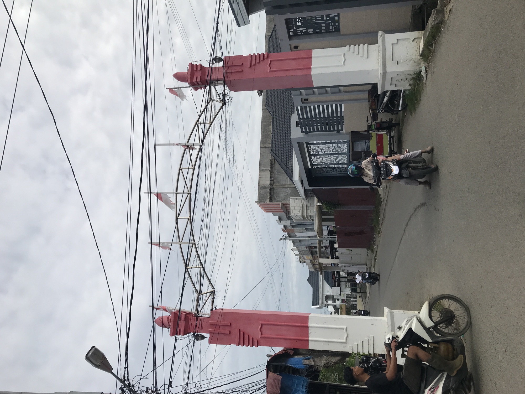 This image shows a street in Lhokseumawe, Indonesia. In the foreground, a large archway spans the street. The archway is supported by two pillars, painted red and white, the colors of the Indonesian flag. The arch itself is made of metal and decorated with small Indonesian flags. A dense tangle of electrical wires obscures the sky behind the archway.
On the right side of the street, there are several buildings with varying architectural styles. People are riding motorcycles on the street, and some are parked along the side. The street is paved and appears to be in a residential or mixed-use area. The sky is overcast, and the overall lighting suggests it is daytime.