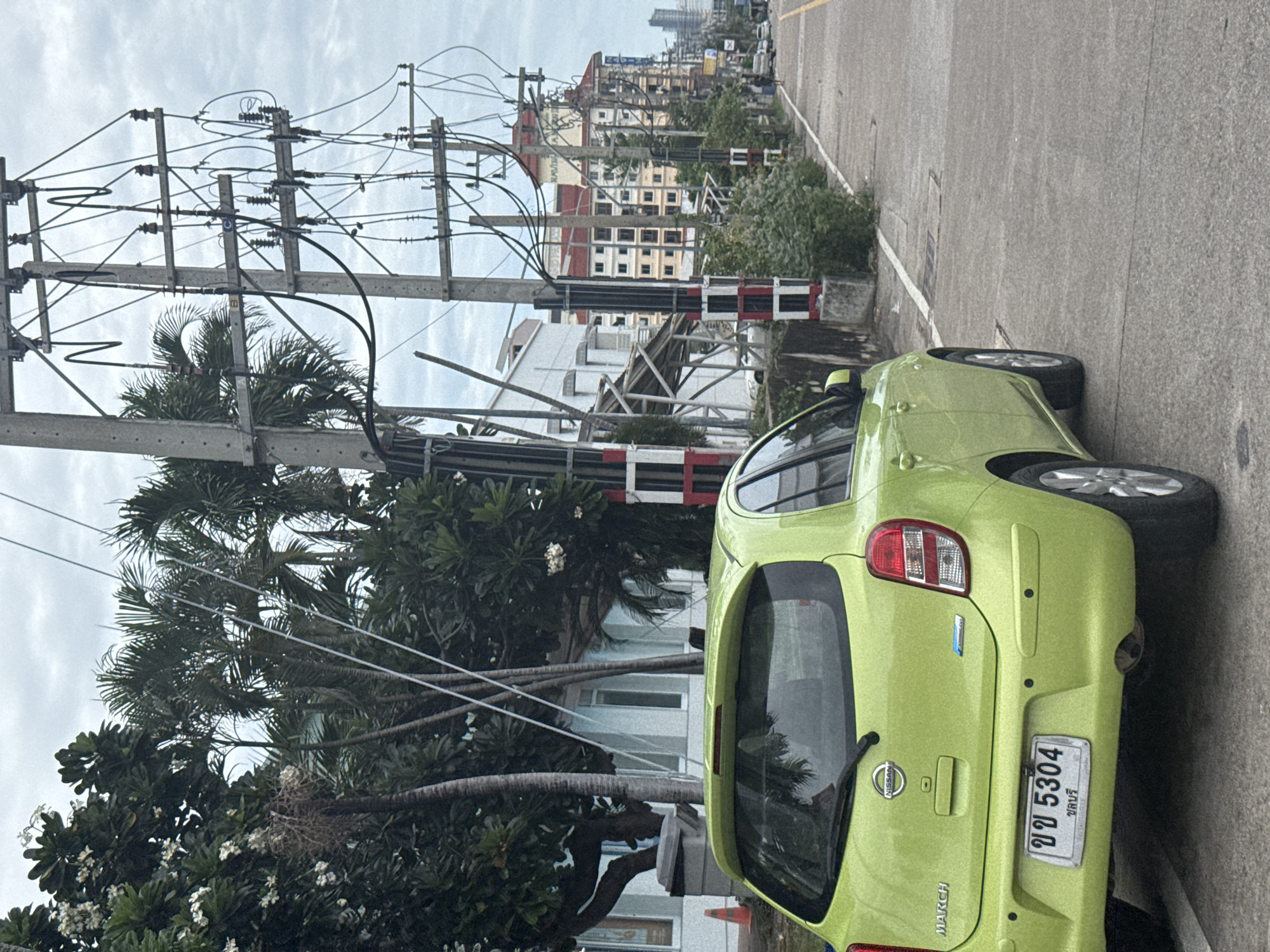 The image shows a bright green Nissan March car parked on the side of a street in Pattaya City, Thailand. The car has a license plate with Thai characters and numbers. To the left of the car are trees with white flowers and palm trees, as well as a light-colored building with windows. To the right of the car is a concrete road with a white line marking the edge, and a row of utility poles with numerous electrical wires. Beyond the utility poles, there are several multi-story buildings, some with red roofs, characteristic of urban development in Pattaya. The sky is overcast. In the distance, more buildings and a hazy horizon are visible.