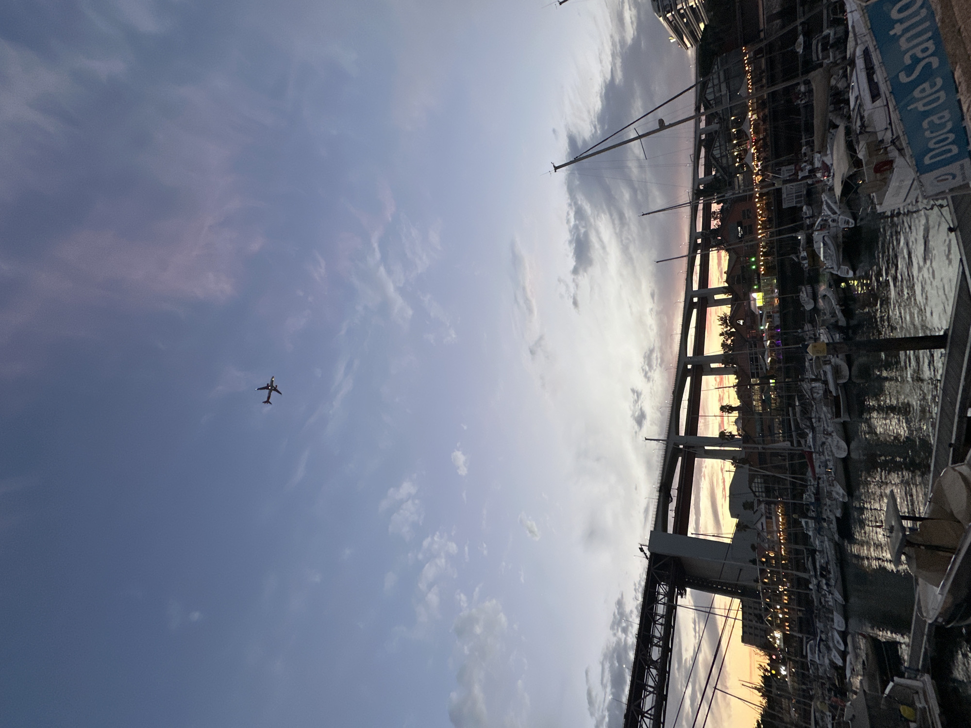 This media file depicts a marina at dusk, likely in Lisbon, Portugal. The scene is dominated by a dramatic sky with a mix of soft pinks and grays, indicating either sunset or sunrise.
The primary subjects are numerous sailboats docked in the water, their masts creating a complex network against the sky. A large, modern bridge structure spans across the marina in the background, its lights beginning to glow as darkness descends. On the right side of the image, a blue sign with white text is visible, reading "Doca de Santo Amaro," which is a well-known marina in Lisbon.
In the vast expanse of the sky on the left side of the frame, a silhouetted airplane can be seen flying, its wings outstretched. The overall mood is serene and picturesque, with the soft lighting and the quiet presence of the moored boats suggesting a peaceful end to the day. The reflection of the lights from the buildings and boats shimmers on the water's surface, adding to the tranquil ambiance.