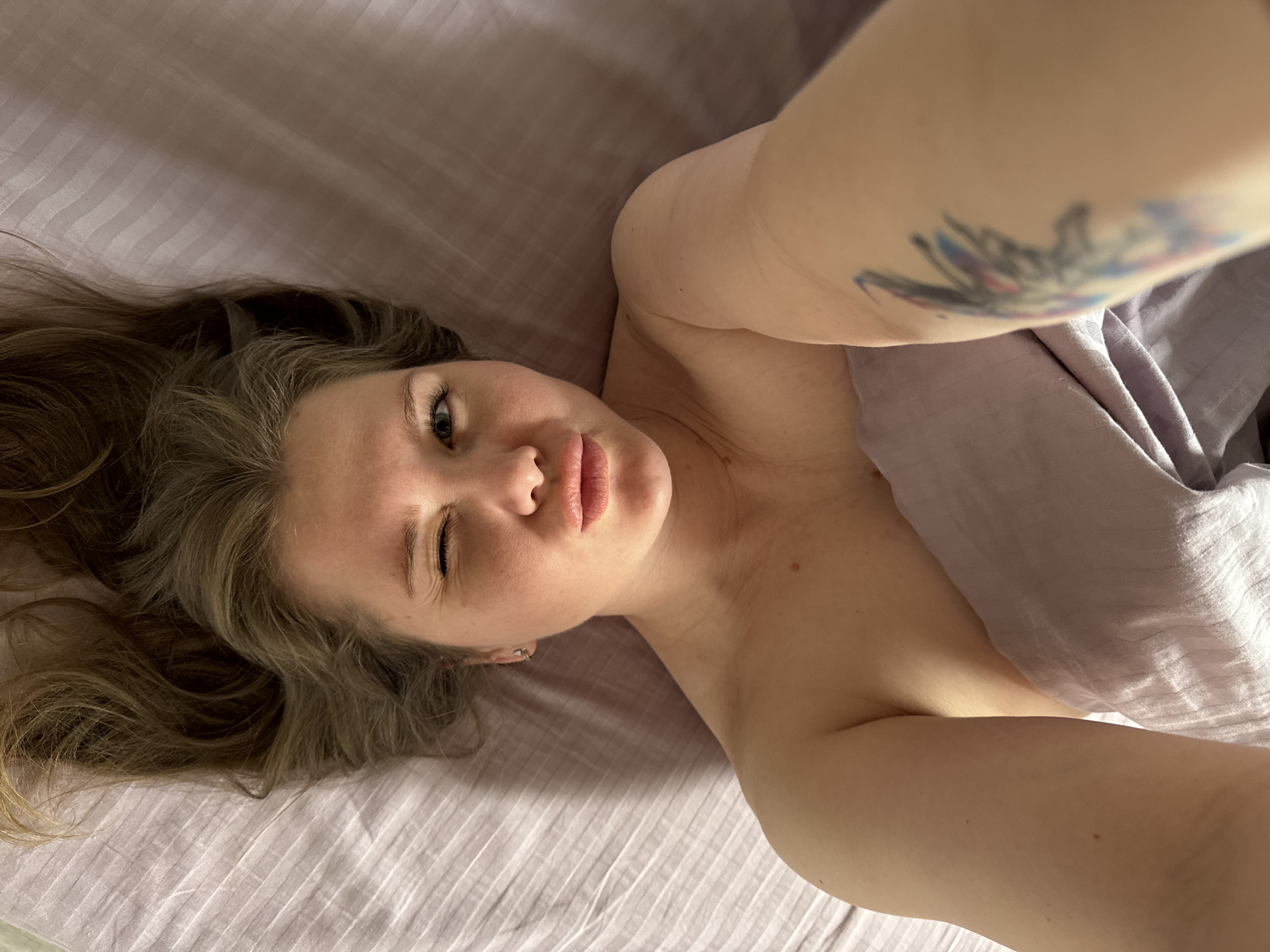 A woman is lying in bed, partially covered by a light purple sheet. She has light brown hair spread across the pale purple bedding and is winking her left eye while looking directly at the camera. Her right arm is raised, revealing a tattoo on her upper arm. The background is out of focus, showing only the bed and the surrounding area. The location is Ферма, Russia.