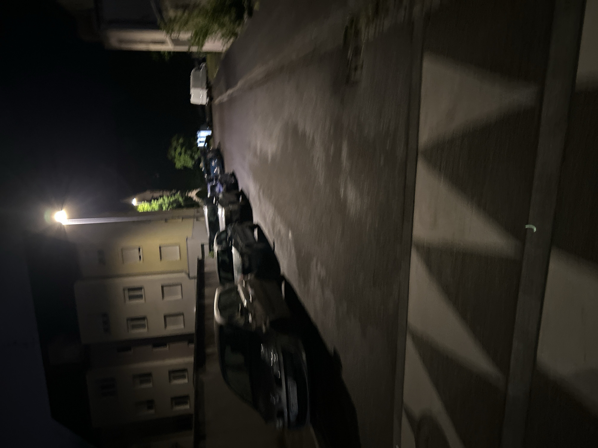 An oblique, low-angle, nighttime view of a street in Strasbourg, France, shows parked cars along the left side of the road. The scene is illuminated by streetlights. A building with multiple windows is visible on the left. A crosswalk is visible on the right side of the street.