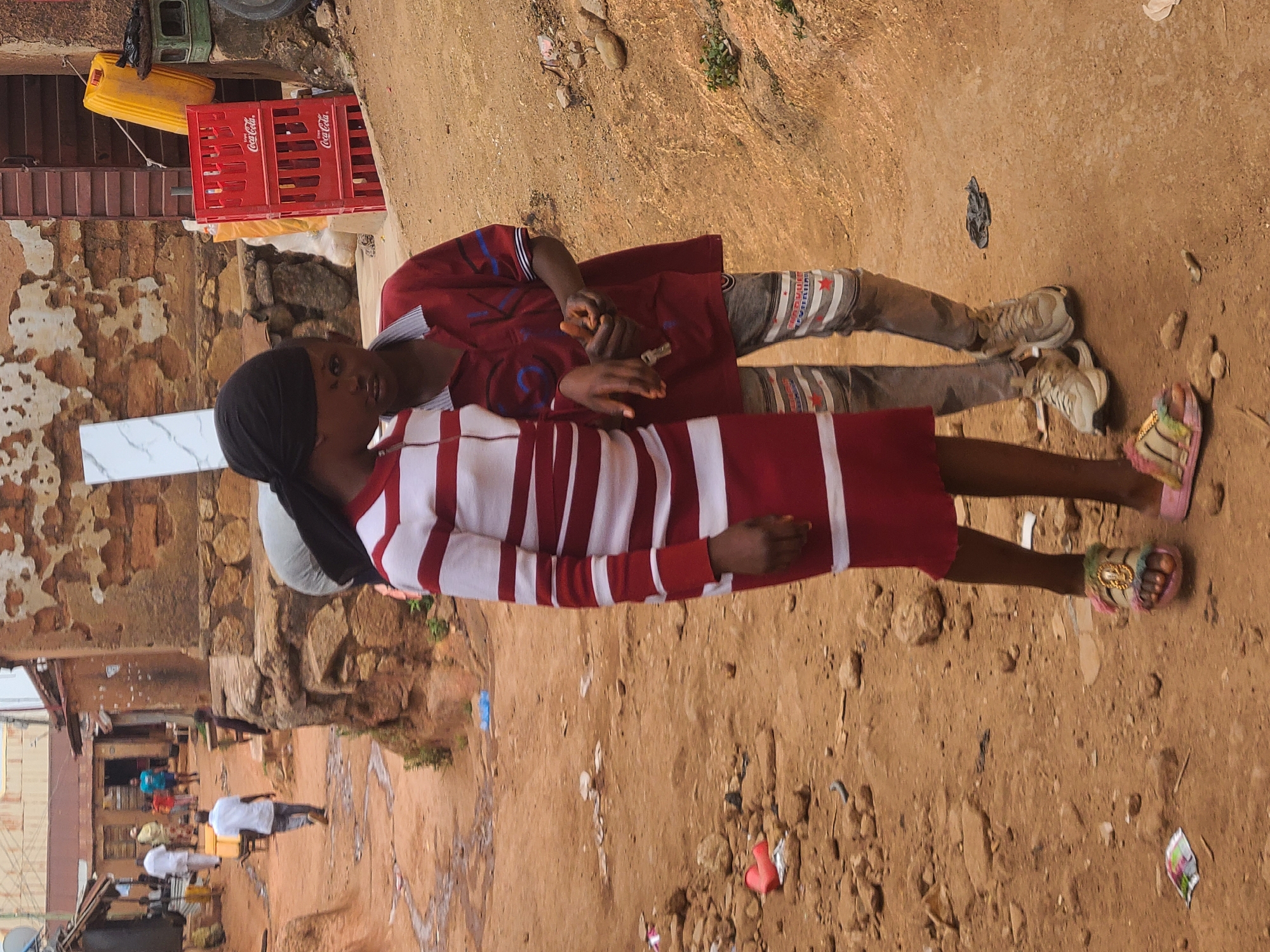 Two children lie on the ground. The child on the left is wearing a white and red striped dress with a black head covering. The child on the right wears a red shirt and patterned gray pants. Both are barefoot. The ground is brown dirt and scattered debris. Background includes buildings, red Coca-Cola crates, and a yellow container. The location is Jos, Nigeria.