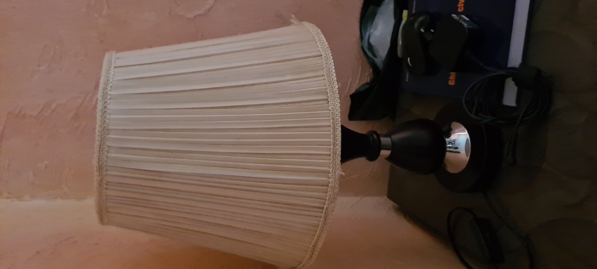 The image shows a lamp with a pleated cream-colored lampshade. The base of the lamp appears to be black with a reflective silver accent. To the right of the lamp, several objects are visible, including what looks like a computer mouse, a black notebook, and a coiled cable. The background is a textured pink wall. The lighting in the image is dim, suggesting it might be nighttime or an indoor setting with low light. The overall scene appears to be a personal workspace or living area. There is no discernible activity taking place, and no individuals are present. The focus is on the stationary objects. Some text is visible on the spine of the black notebook, but it is too blurry to read.