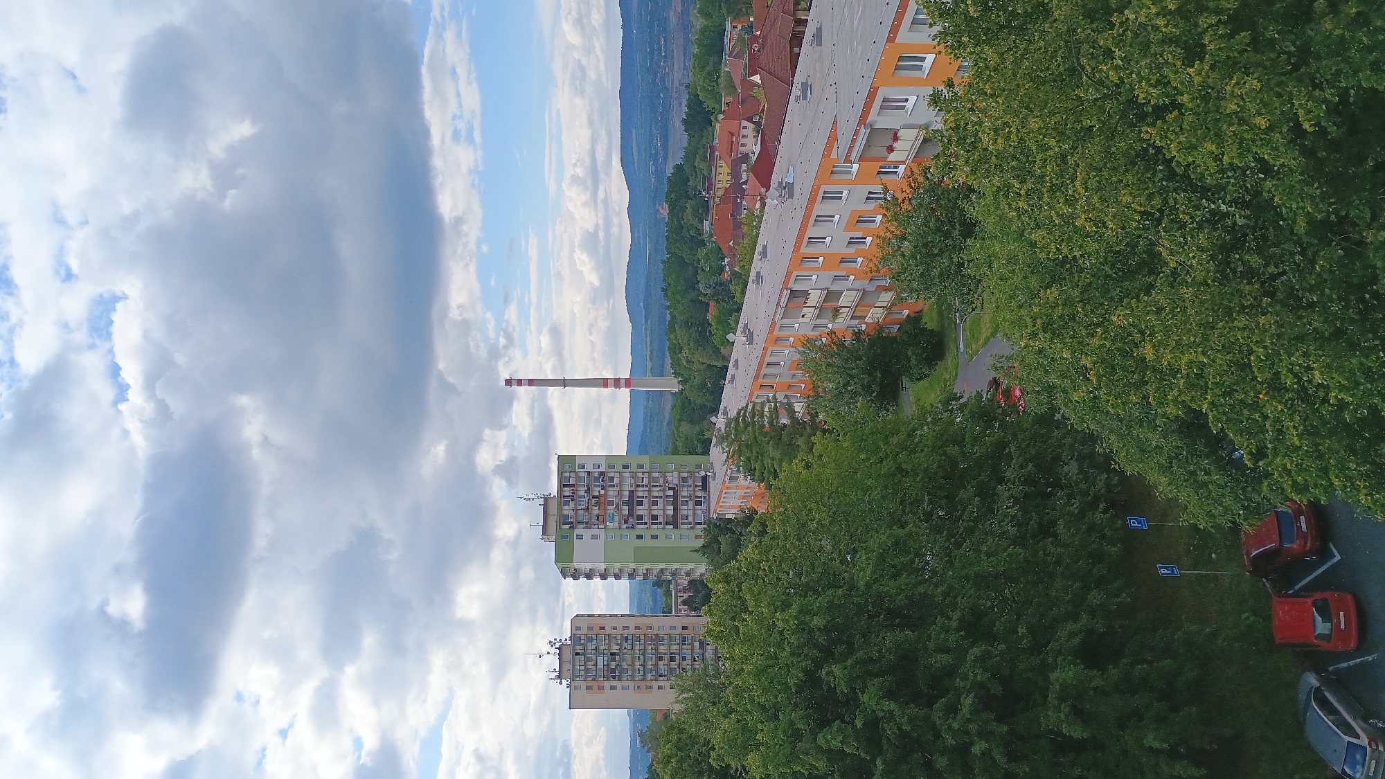 An aerial view captures a residential area in Sokolov, Czechia, under a partly cloudy sky. Dominating the scene are several multi-story apartment buildings. Two tall, cream and green apartment blocks stand side-by-side in the mid-ground, featuring numerous balconies. To their right, a long, low-rise apartment building with orange and gray facades stretches across the frame. In the distance, a tall, red and white striped industrial chimney pierces the sky, characteristic of Sokolov's industrial landscape. The foreground and mid-ground are densely populated with lush green trees, suggesting a temperate climate. A parking area is visible at the bottom left, with several cars parked, including a silver sedan, a red hatchback, and a dark red SUV. Blue parking signs are also present. The overall impression is of a typical urban residential environment integrated with significant green spaces.