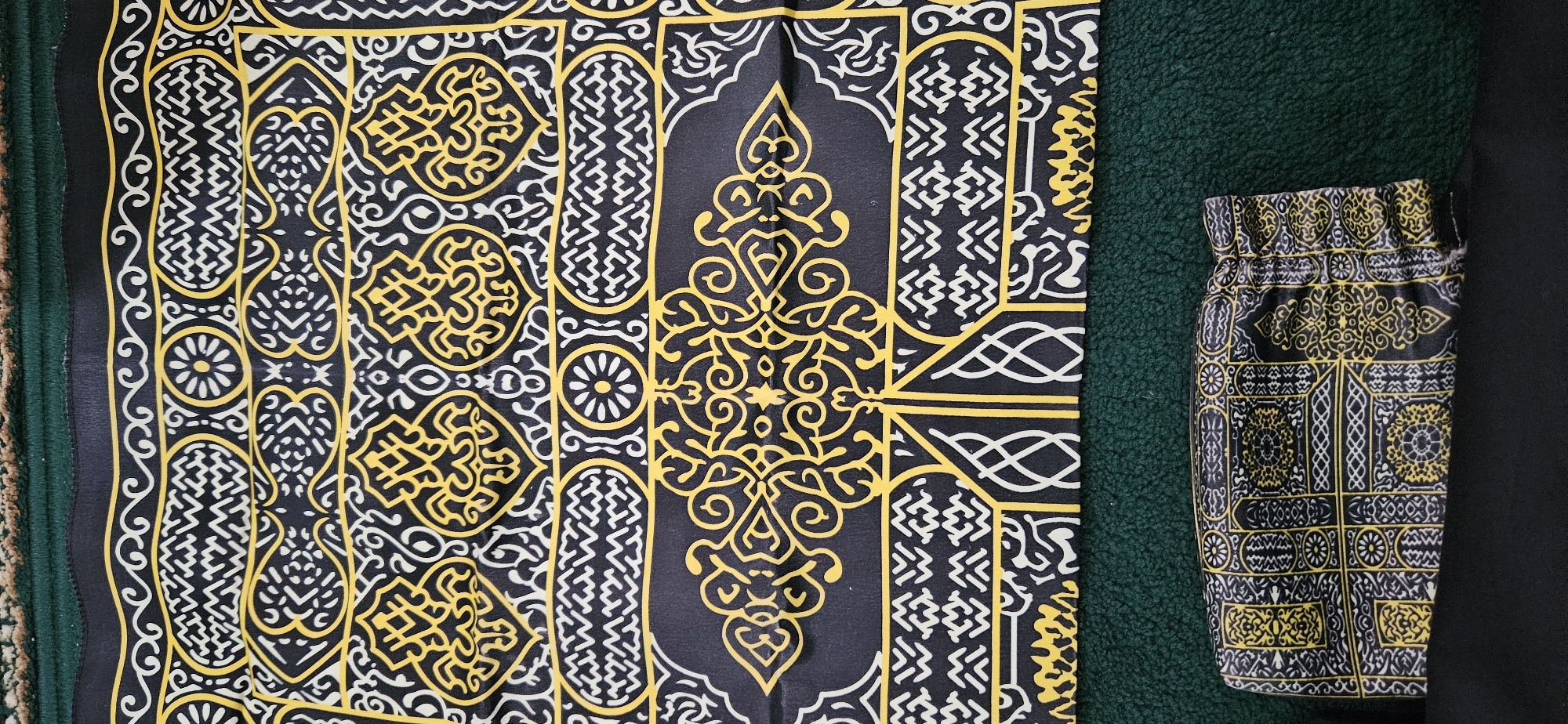 This is a top-down, medium shot of a prayer mat. The mat is black with intricate yellow and white geometric and floral patterns. The patterns appear to be Islamic in design. The mat is folded and lies on top of a green carpet or rug. The background of the image is a dark green, textured material. Below the prayer mat, a small, folded black pouch with a similar pattern to the mat is visible. The scene is indoors, and there are no people or animals present. The lighting is diffused and even, suggesting an indoor setting with ambient light. The image is sharp and clear, allowing for detailed observation of the fabric's texture and the design's intricacies. The location is unspecified, but the subject matter (a prayer mat) suggests a religious context, possibly a home or a mosque.