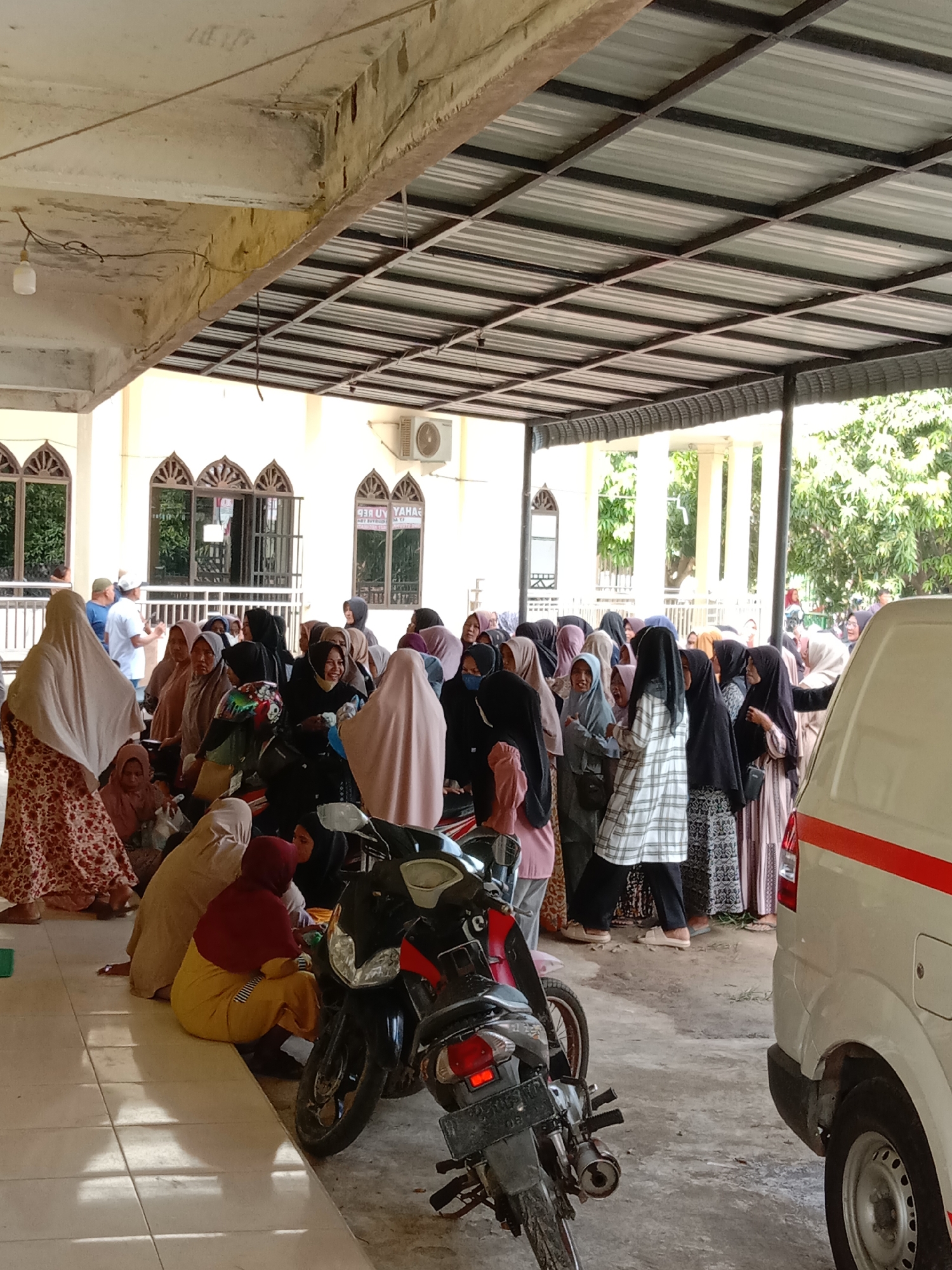 The scene shows a gathering of people, predominantly women wearing hijabs, under a covered area adjacent to a building with arched windows, possibly a mosque, in Lhokseumawe, Indonesia. Some women are sitting on the floor, while others are standing. There are parked motorcycles and a white vehicle with a red stripe on the right side of the frame. The area appears to be an outdoor waiting area. The location is likely near a public building in Lhokseumawe, Indonesia.