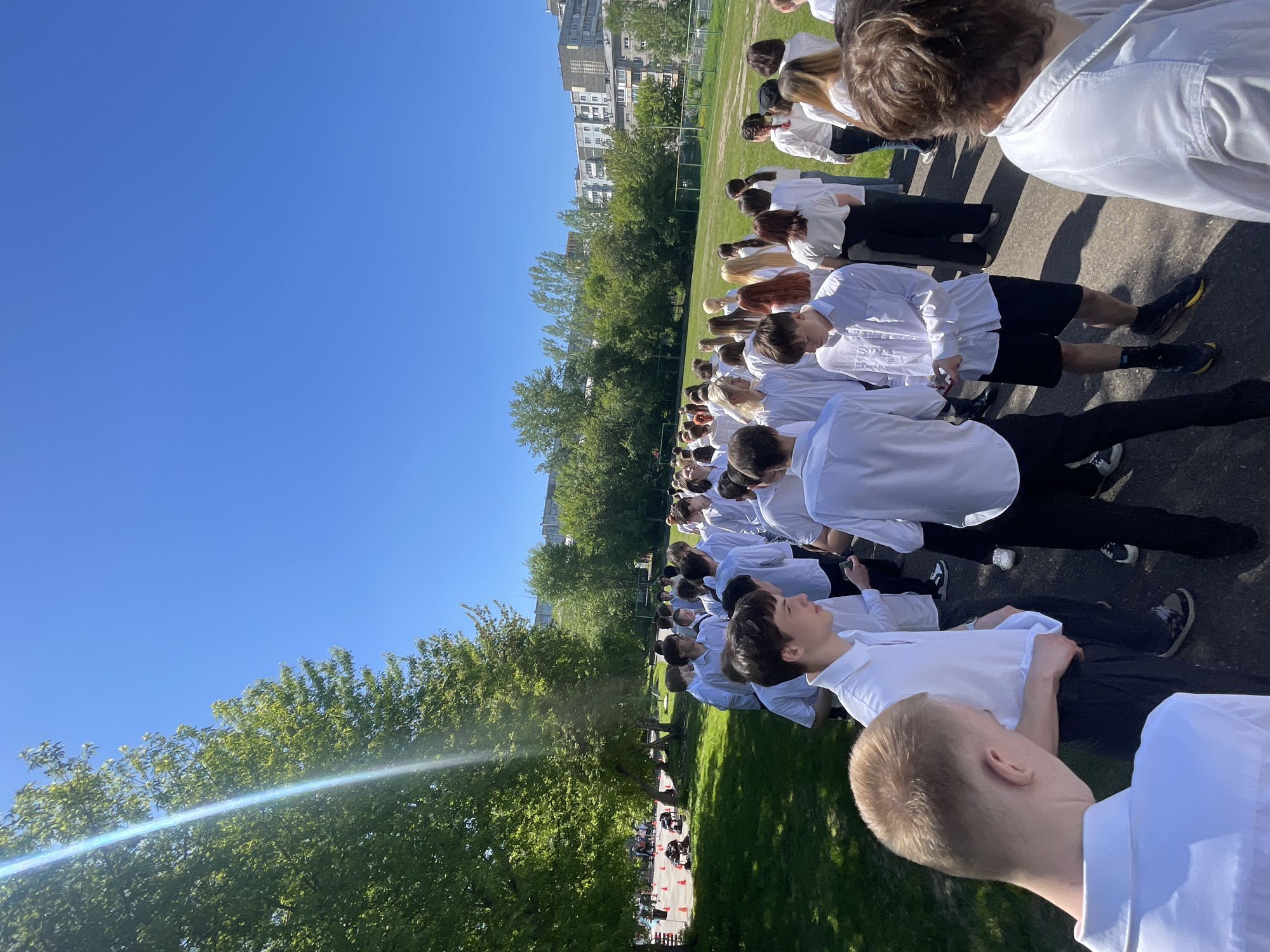 The image depicts an outdoor gathering of a large group of young individuals, likely teenagers or young adults, on a bright sunny day in Krasnoyarsk, Russia. The subjects are uniformly dressed in white shirts and dark trousers or shorts, arranged in rows on a paved or asphalt surface. Many are seen from behind or in profile, looking forward or upward.
The setting includes a wide expanse of clear blue sky overhead, a vibrant green grassy field adjacent to the paved area, and numerous tall green trees. In the background, multi-story urban buildings are visible, suggesting a city or suburban location. On the far side of the grassy field, there are orange cones and possibly small vehicles, hinting at an organized activity, course, or competition.
The strong shadows cast by the people indicate bright sunlight, suggesting the time of day is midday or early afternoon. The weather is clear and sunny. The group's uniform attire and orderly arrangement imply an organized event, such as a school assembly, sporting event, or performance. No visible text is present in the image.