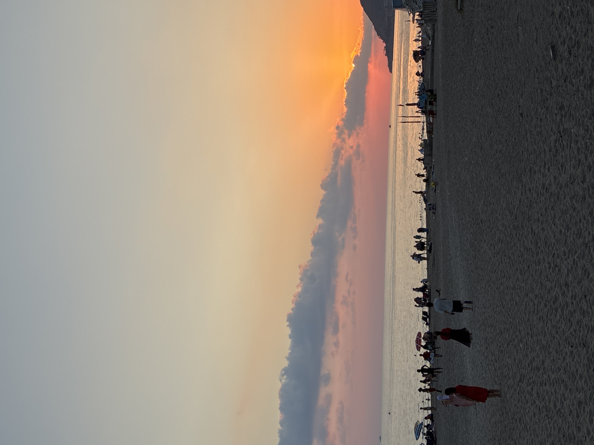 An image taken at sunset on Cleopatra Beach in Alanya, Turkey, captures the serene atmosphere of a public beach. The sky is a gradient of soft oranges and pinks, with a few scattered clouds adding texture to the horizon. The sun, partially obscured by clouds and a distant headland, casts a warm glow across the tranquil sea.
On the sandy beach, numerous people are visible enjoying the evening. Some are walking along the shoreline, while others are gathered in groups, possibly socializing or resting. Several beach umbrellas and chairs are set up, indicating areas for relaxation. The water appears calm, with a few small boats or buoys visible in the distance. The overall scene depicts a typical evening at a popular tourist destination, with a mix of leisure activities and natural beauty.