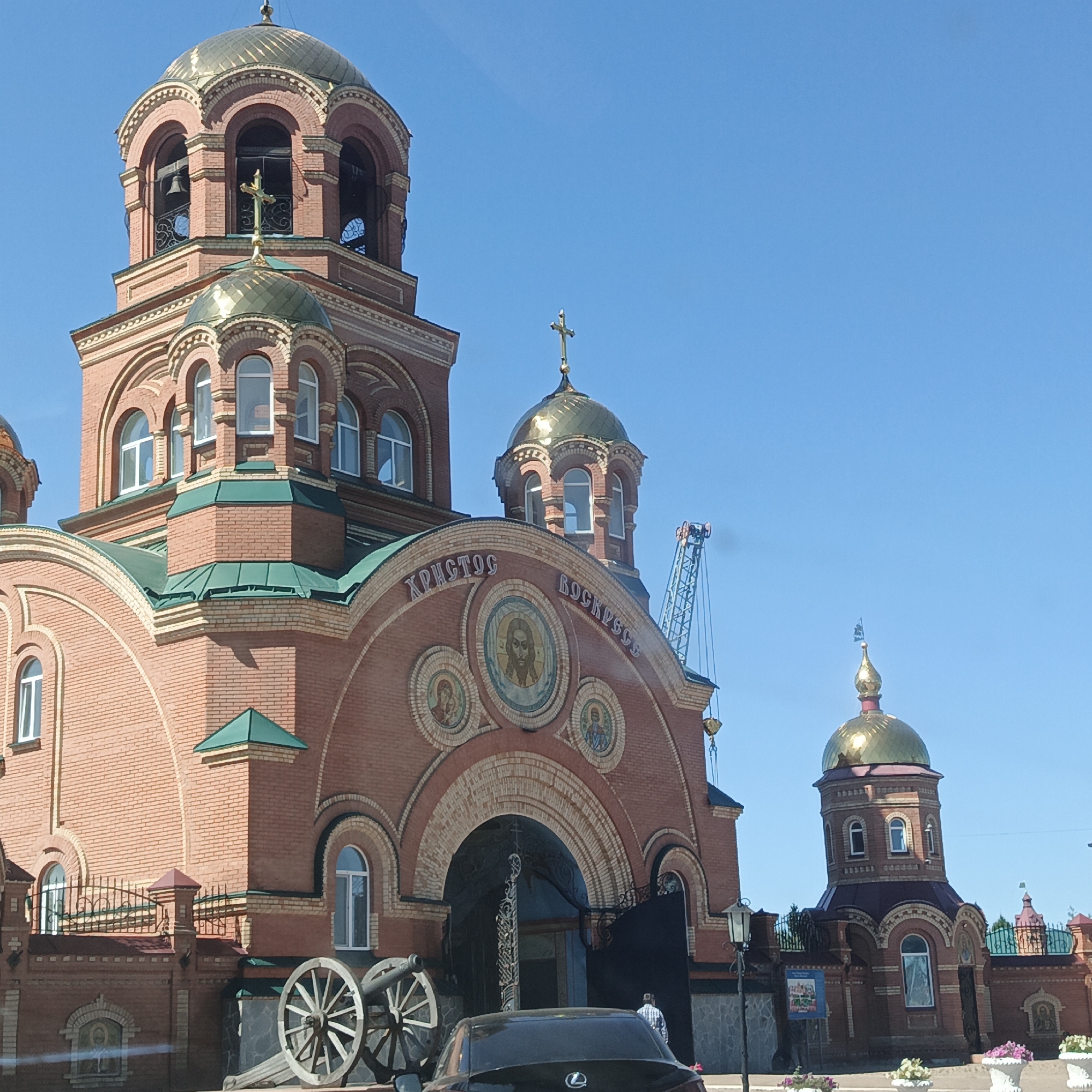 The image captures the Church of St. Nicholas the Wonderworker in Saraktash, Russia. The predominantly brick church features golden domes and crosses, characteristic of Orthodox architecture. The building displays intricate brickwork with yellow decorative elements and green tiled roofs. On the main facade, above the entrance, is a large mosaic depicting Jesus Christ, flanked by smaller mosaics and inscriptions. To the left of the entrance, two historical cannons are displayed. A dark-colored car is parked in front of the church, and a few people are visible near the entrance. In the background, a construction crane is present, suggesting ongoing work or renovation. The sky is a clear, bright blue.