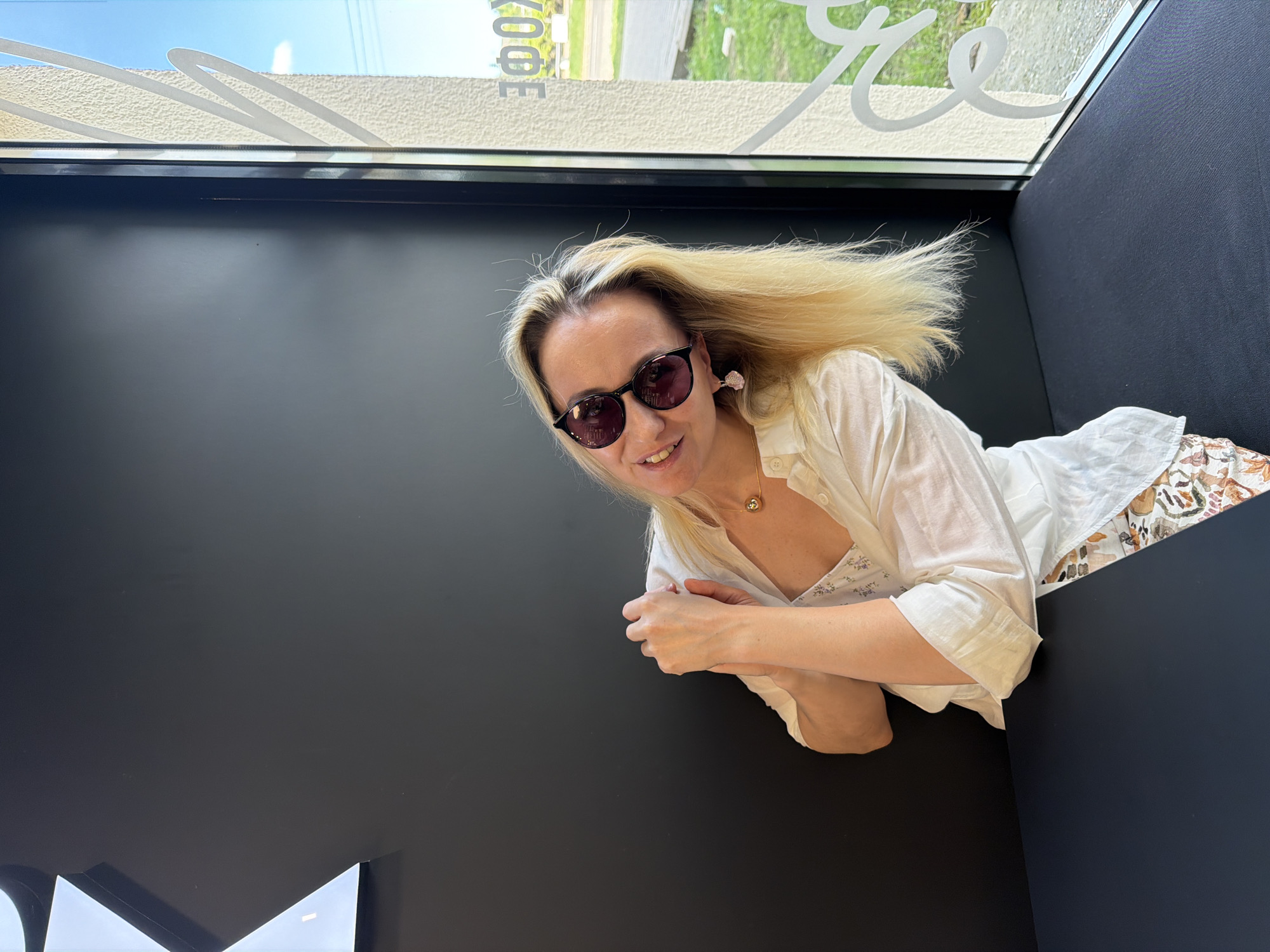 A woman with blond hair and sunglasses is visible from an elevated perspective. She is wearing a white shirt and floral patterned skirt. She is resting her arms on a dark, possibly black, surface. In the background, through a window, vegetation and a building are visible. The reflection in the window also shows lettering, potentially indicating the location of the scene as being in Izhevsk, Russia. The lighting appears to be bright, suggesting daytime.
