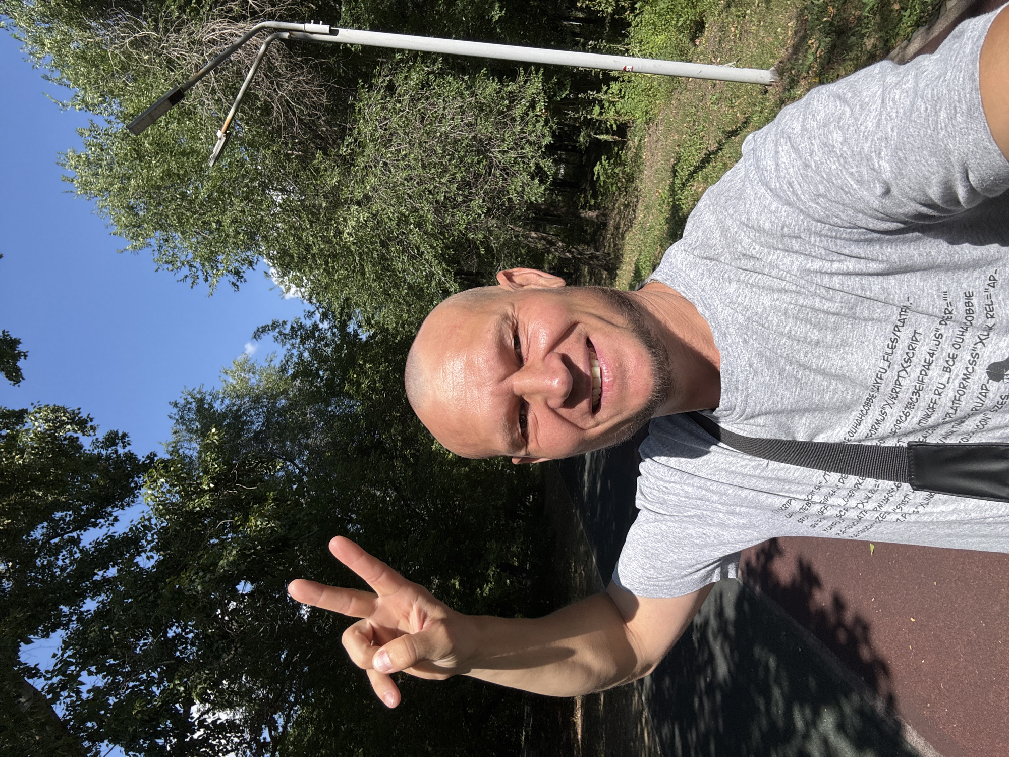 A bald, Caucasian man in his 40s, with a goatee, is taking a selfie outdoors in Oskemen, Kazakhstan. He is wearing a grey t-shirt with text printed on it and a black crossbody bag. His left arm is raised, and he is making a peace sign with his fingers. He is smiling at the camera. The background consists of trees and a blue sky. There is a street light on the right side of the image. The ground is a mixture of dark grey asphalt and red surface, possibly a running track. The scene appears to be a park or recreational area within the city.
