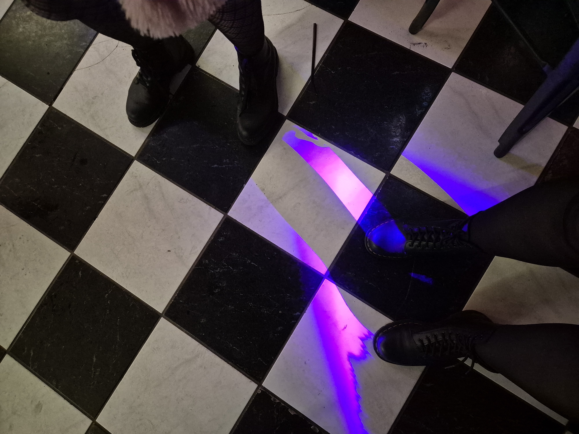 The image shows a checkered black and white floor with two people standing on it. One person on the left is wearing black fishnet stockings and black shoes. The other person on the right is wearing black pants and black boots. A purple and pink light beam shines across the floor between the two people. A black stick is also present on the floor. A chair leg is visible in the top right. The location is likely within West Suffolk, United Kingdom, given the instructions.