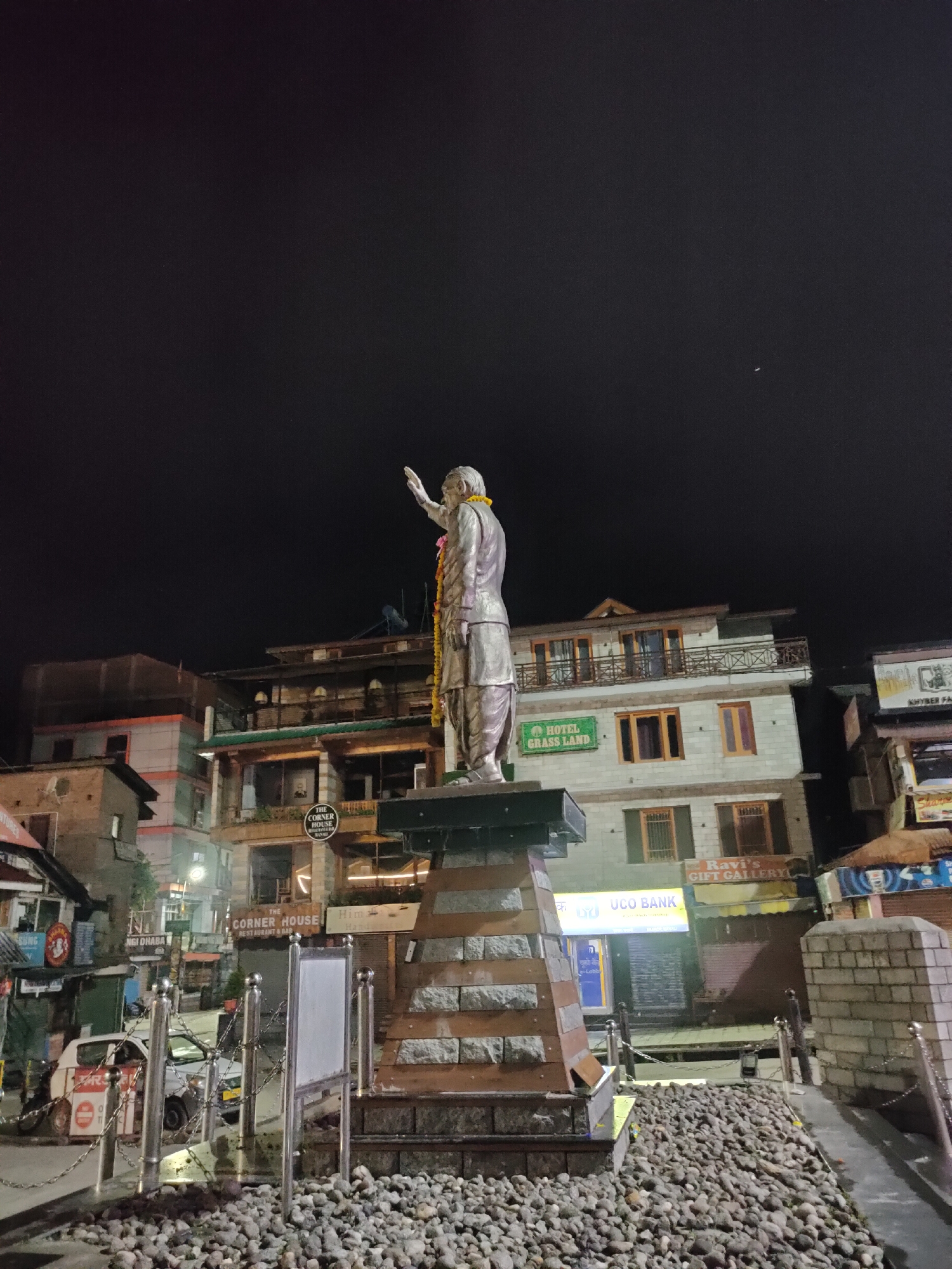 The image captures a statue of a man, adorned with a yellow garland, standing tall at night in the city of Manali, India. The statue is situated on a multi-tiered pedestal made of stone and wood. Behind the statue, a street scene unfolds with buildings featuring multiple stories, balconies, and illuminated windows. Signs visible on the buildings include "HOTEL GRASS LAND," "UCO BANK," and "Ravi's GIFT GALLERY." A white car is parked on the left side of the street. The foreground is covered with small, gray stones. The sky above is dark, indicating nighttime.