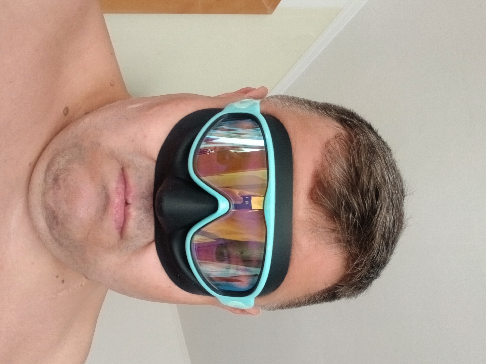 A close-up, head-on, waist-up shot shows a middle-aged man wearing black swimming goggles with teal frames and iridescent lenses. He has short, graying hair and is shirtless. The background is a plain, light-colored wall, with a hint of a doorway or furniture to the left. The lighting is even, suggesting an indoor setting. The man's face is partially obscured by the goggles, which cover his eyes and the bridge of his nose. His mouth is visible, with his lips slightly parted. The image appears to have been taken in a residential interior, possibly a bedroom or bathroom, in Pfarrkirchen, Germany.