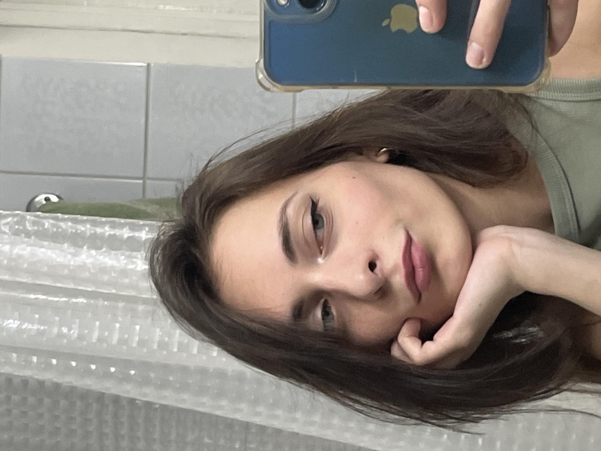 A young woman is taking a selfie in what appears to be a bathroom. She is holding an iPhone with a blue case. The background includes tiled walls, a shower curtain, and a metallic faucet. The woman has brown hair and is wearing a green top. She is resting her head on her hand and looking at the camera. There is no discernible evidence to connect the image specifically to the city of Krasnoyarsk, Russia.