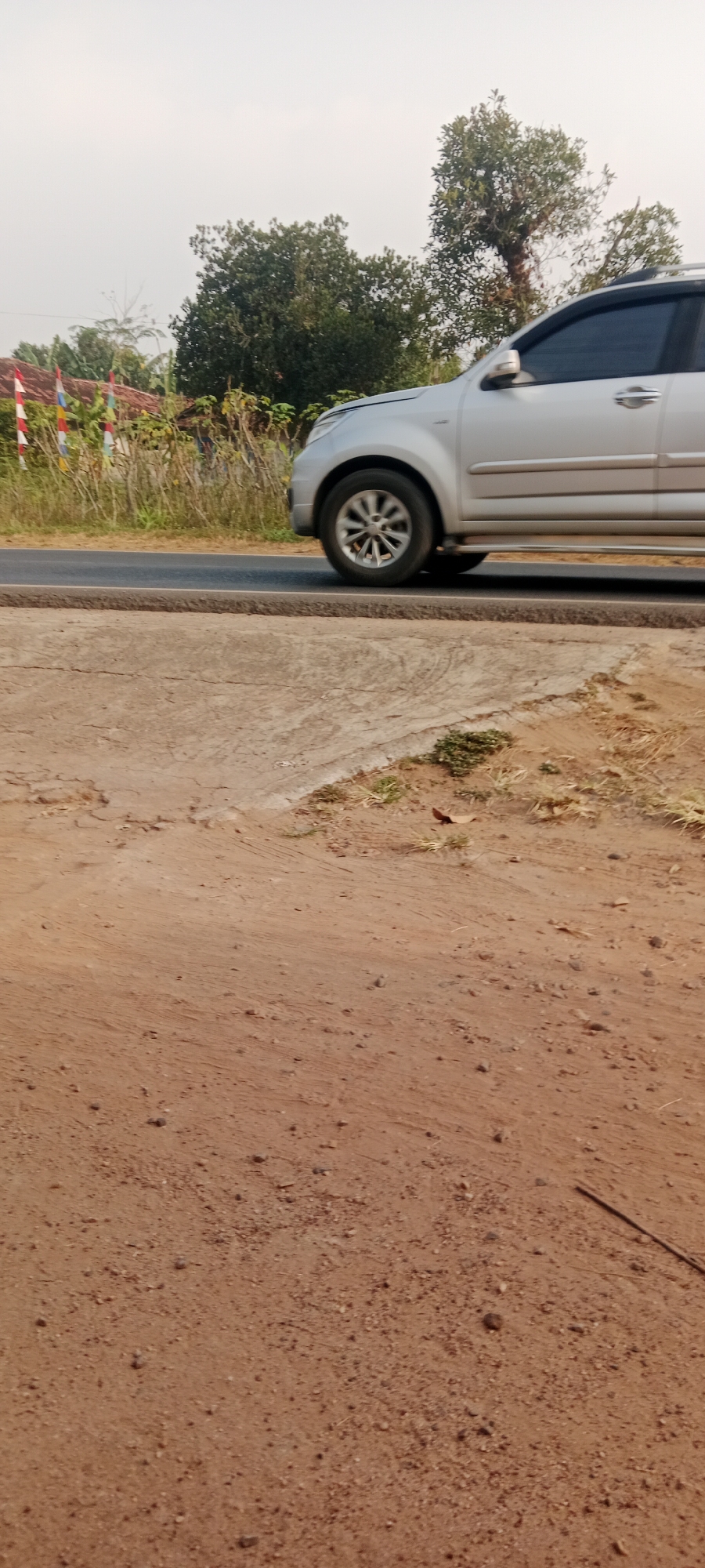 The image, captured in Gondang Rejo, Indonesia, presents a roadside scene. A silver SUV is shown in motion on a paved road. To the left of the road, there's a stretch of dry grass and vegetation, with a few colorful flags planted in the ground. Behind the vegetation, a building with a reddish roof is partially visible. The sky above is overcast. The foreground of the image consists of a dirt path with scattered pebbles. The overall environment suggests a rural or semi-rural setting.