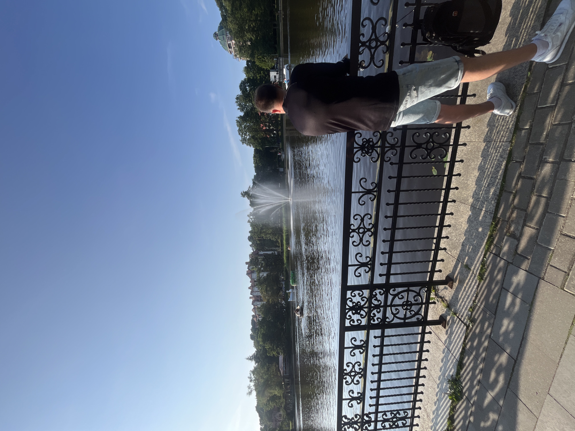 A man stands at a black metal railing overlooking a calm lake with a fountain spraying water in the distance. He is facing away from the camera, wearing a black long-sleeved shirt and light blue denim shorts. The man has white socks and white sneakers. A black backpack hangs on the railing to his right. The lake is surrounded by green trees and buildings, including a domed building on the right side of the frame. On the lake, there are several pedal boats, including one shaped like a swan and another that is green. The ground in front of the railing is paved with light-colored bricks, and there are shadows cast by the railing on the pavement. The sky is clear and blue. The scene appears to be in Svetlogorsk, Russia, based on the architectural style and the presence of a lake with recreational boats.