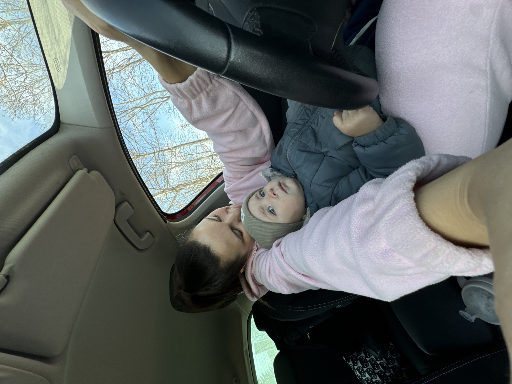 The image depicts a person holding a baby inside a vehicle. The person is wearing a pink top. The baby is wearing a gray jacket and hat. The interior of the vehicle is visible, including the steering wheel, the dashboard, and the roof. The background shows trees and a blue sky. The location of the image is Unknown, Unknown.