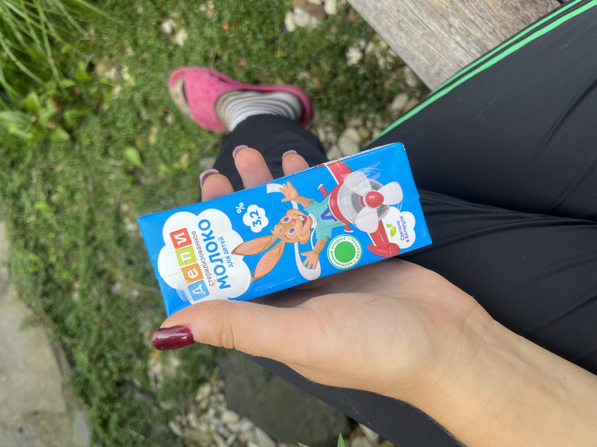 In an outdoor setting with green foliage and a wooden surface, a person's hand holds a blue juice box. The juice box displays a cartoon rabbit and text in Cyrillic. The person is wearing black pants with green stripes. A pink slipper is visible on their foot in the background. The image appears to be taken in the Дзямідавіцкі сельскі Савет, Belarus.