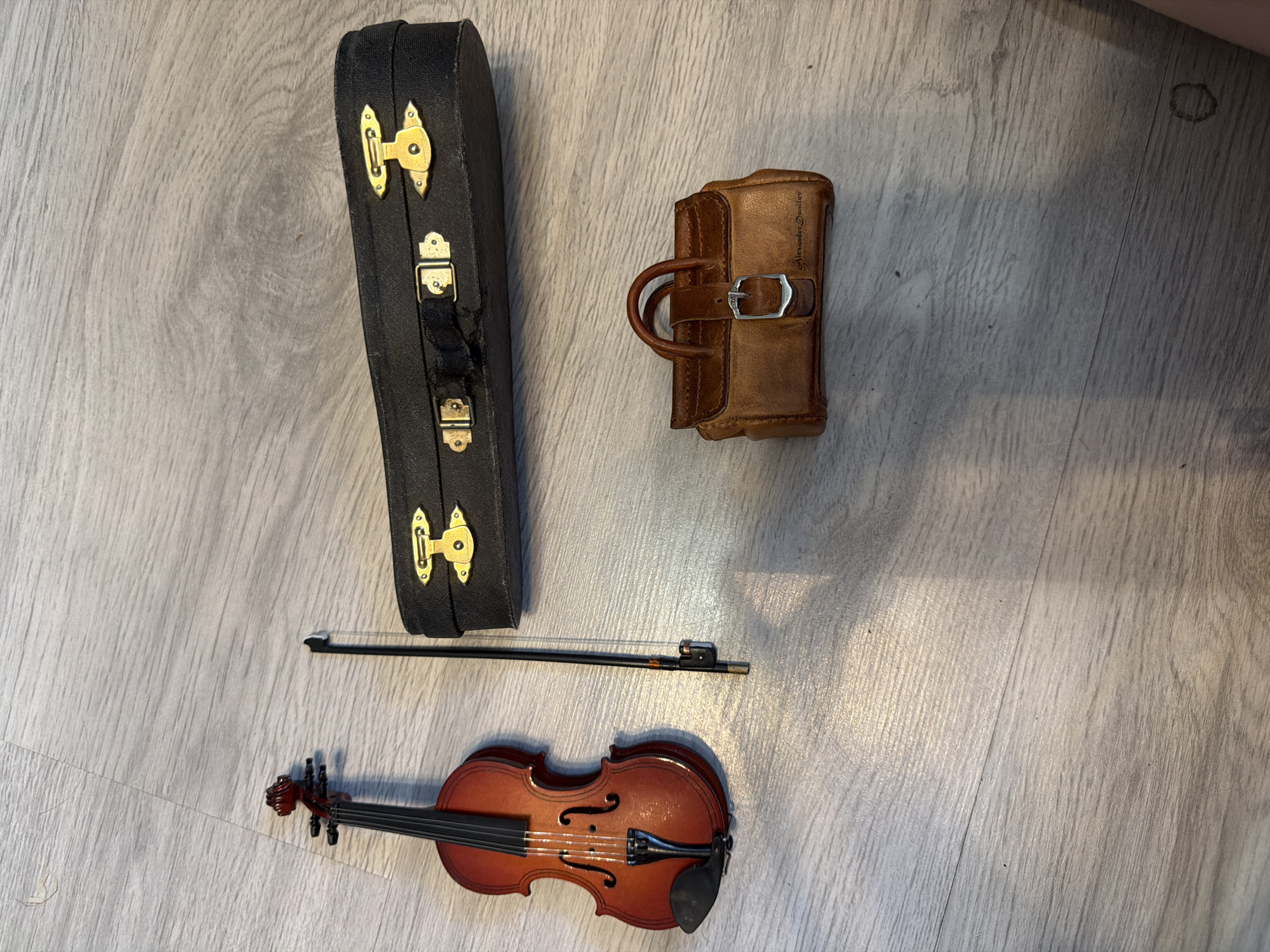 A violin and bow are laid out on a wooden floor next to a black violin case and a small brown leather bag. The violin is made of wood with a reddish-brown finish. The bow is black and has a silver metal frog. The violin case is rectangular with gold latches. The leather bag is small and appears to be a miniature handbag with a buckle closure. The floor is a light gray wood grain pattern. The scene is well-lit, with some sunlight casting shadows on the floor. The objects are arranged neatly, suggesting they may be part of a display or collection. There are no people visible in the image.
