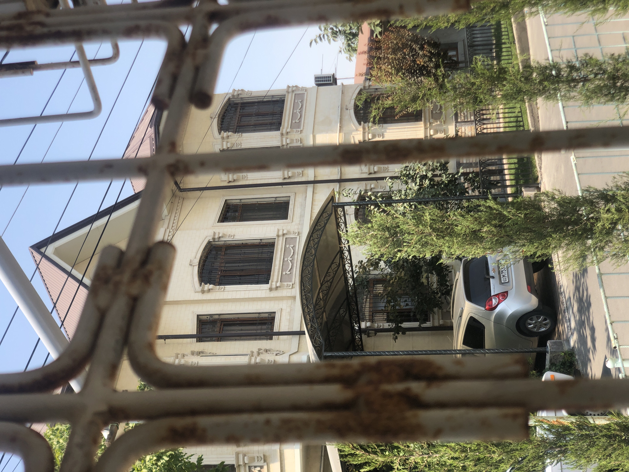 The image captures a street view in Tashkent, Uzbekistan, seen through a rusty metal fence. In the foreground, the weathered bars of the fence dominate, obscuring a clear view. Beyond the fence, a two-story house with cream-colored brickwork and decorative architectural details is visible. The house features arched windows with dark grilles on the lower level and rectangular windows with similar grilles on the upper levels. A covered carport with an ornate metal frame extends from the side of the house.
A silver hatchback car, possibly a Chevrolet Spark, is parked under the carport. The car's license plate is partially visible and appears to be in the Uzbek format, reading "01 M753VA". Several evergreen trees and shrubs are planted in front of and around the house, some of which are taller than the car. The sky is clear and blue, suggesting a sunny day. In the background, another house with a reddish-brown roof can be seen, as well as a white air conditioning unit mounted on the side of the cream-colored house. Power lines crisscross the sky above the structures. The overall impression is a residential street scene in Tashkent on a bright, clear day.