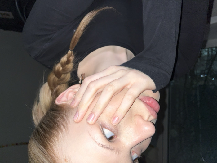 An upside-down perspective captures a person with fair skin, light blonde hair styled in a braid, and a black long-sleeved top. The person's face is partly obscured by their hand, with the fingers spread across the forehead. An earring is visible. A window with blurry reflections is in the background, suggesting an indoor setting.