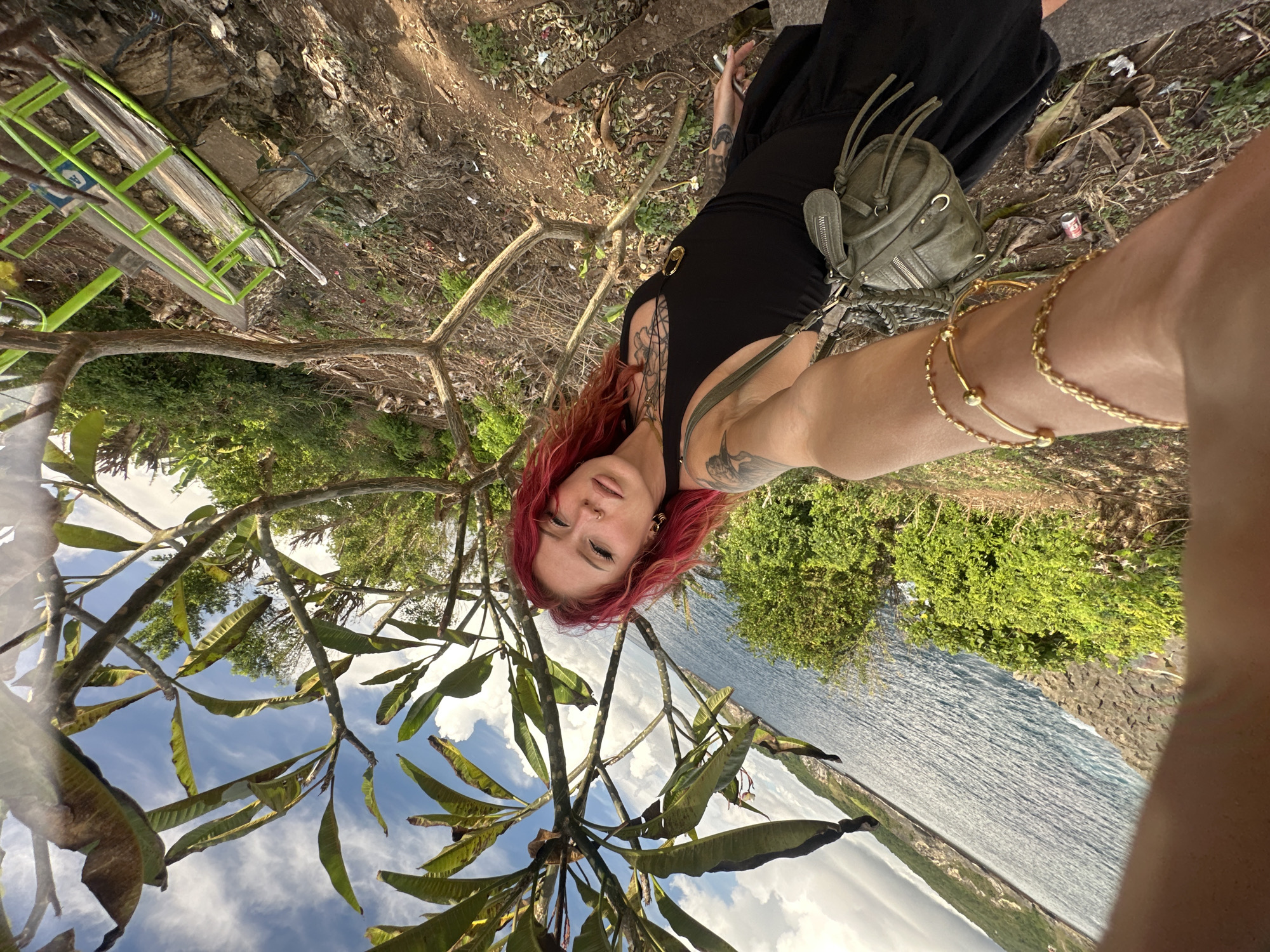 The image shows a woman with red hair and tattoos in Nusa Penida, Indonesia. She is wearing a black dress and a green shoulder bag, and several gold bracelets. The photo is taken from a low angle, looking up through the branches and leaves of a tree towards a view of the ocean, a rocky coastline, and a bright blue sky with clouds. Some litter is present in the background, and part of a green metal structure is visible on the left edge.