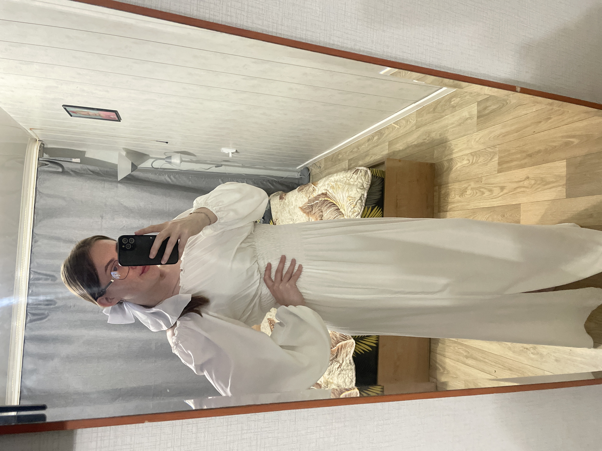 A young woman is taking a mirror selfie in a bedroom. She is wearing a long, white, off-the-shoulder dress with puffy sleeves and a white bow in her hair. She is also wearing glasses. The room has white paneled walls, a gray curtain, a bed with a patterned comforter, and a wooden floor. A floor lamp is visible in the background. The woman's phone, which she is using to take the photo, is visible in her hand. The image appears to have been taken in Сырково, Russia.