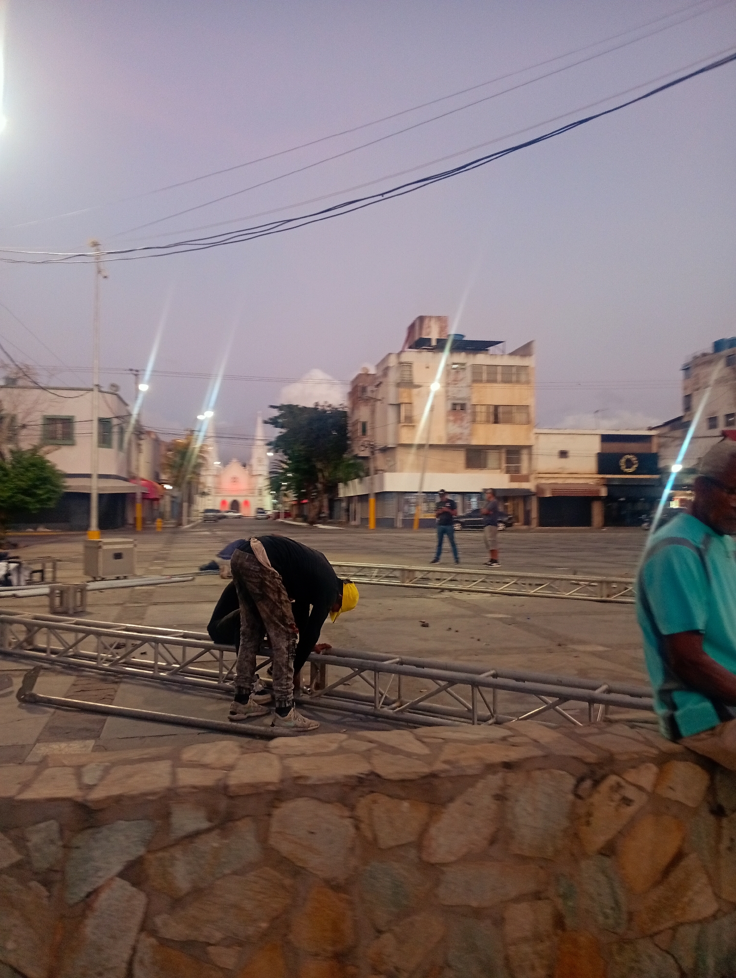 A scene in Juan Griego, Venezuela, shows a public square at dusk. Two men are assembling a metal stage structure, one wearing a yellow hard hat and camouflage-patterned pants. In the background, two other men stand on the open square. A church with white spires is visible. Buildings surround the square. An older man sits on a stone wall in the foreground wearing a teal shirt. Power lines are stretched across the upper part of the scene.